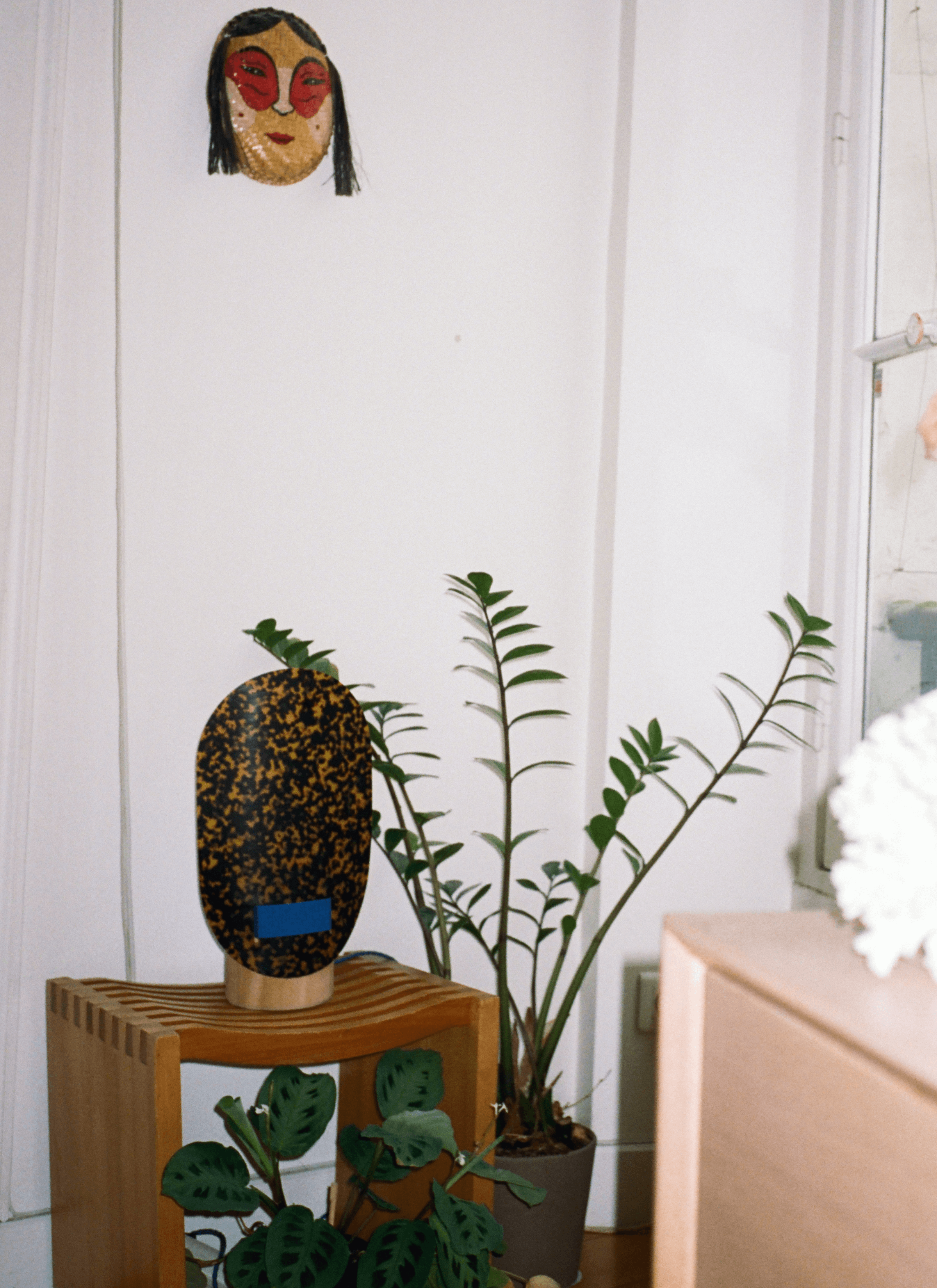In front of a plant, there is a stone-like decoration sitting on a wooden piece of furniture next to a pot of plants. And there is a female kid-like mask hanging on the wall.