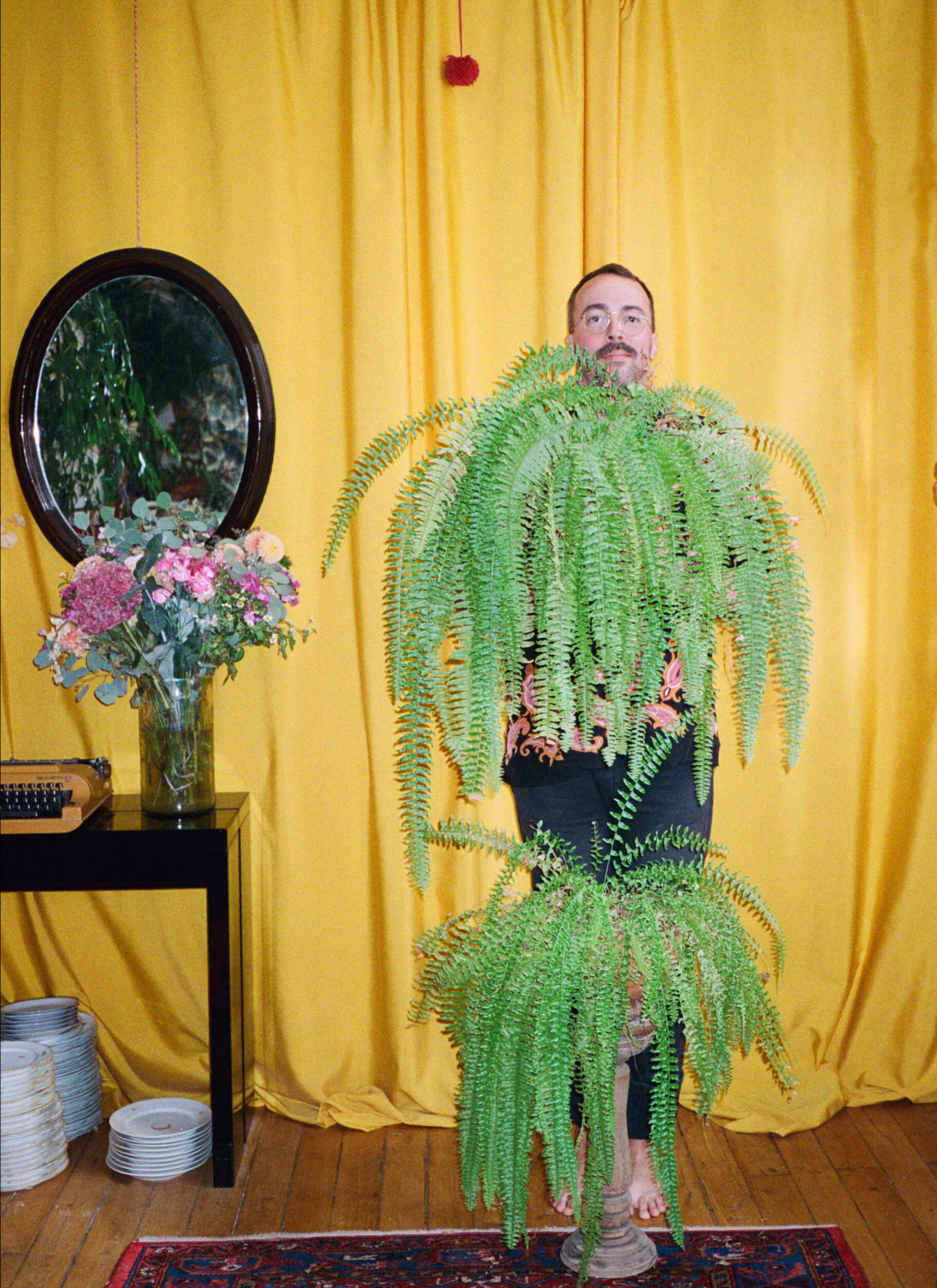 Arnold held a potted plant while another potted plant is in front of him. There are a vase filled with flowers sitting on the desk while an oval shape mirror hung on the yellow curtains in the background.