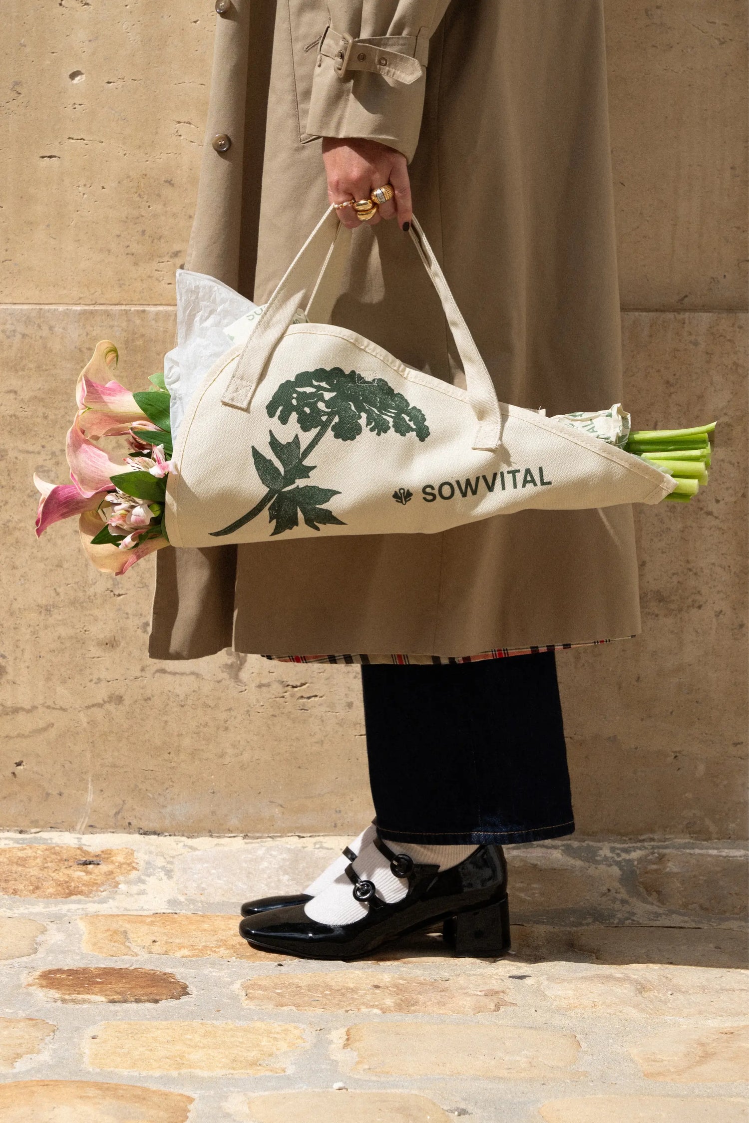 Person holding a white tote bag with green text and floral arrangement against a stone wall.