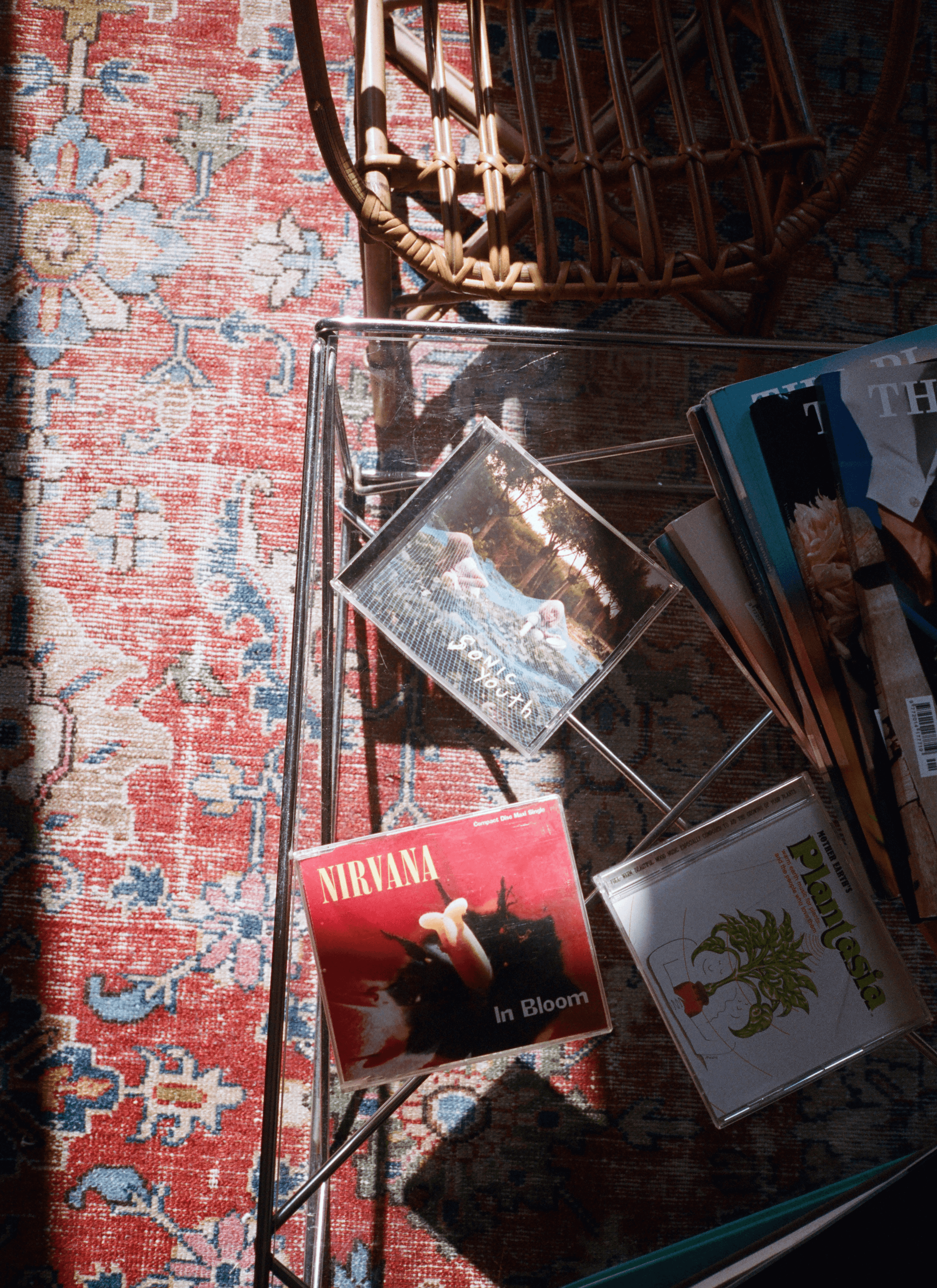 A collection of CDs and magazines on the glass table on a colour-faded carpet next to a bamboo or wooden chair.