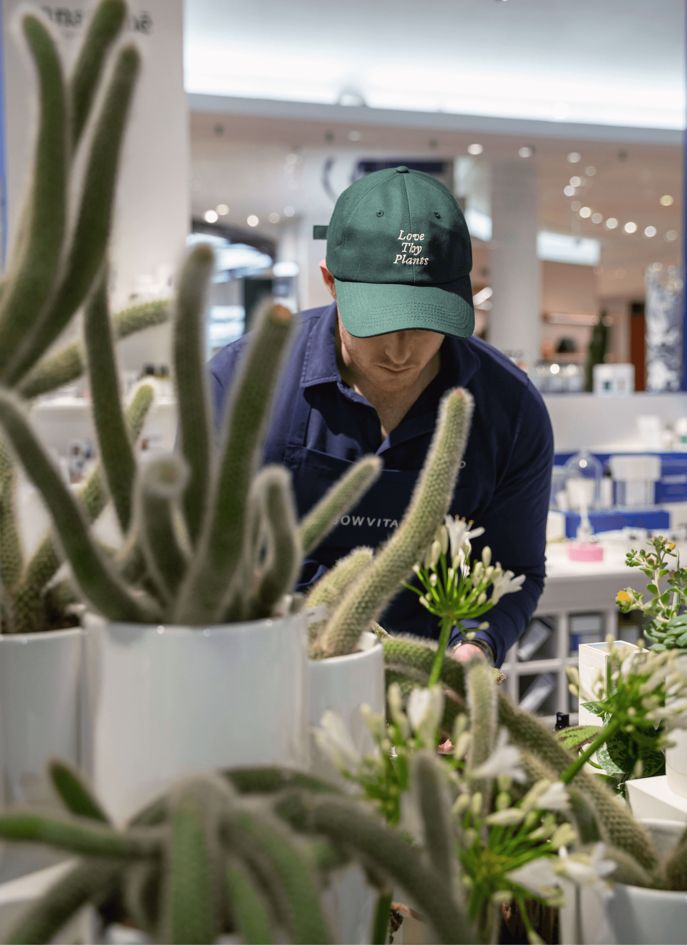 A staff member wearing a green Love Thy Plants cap and a black apron adorned with the Sowvital label tends to a collection of potted plants, flowers, and cacti. With care and attention, they nurture the botanical display, ensuring each plant thrives in its environment.