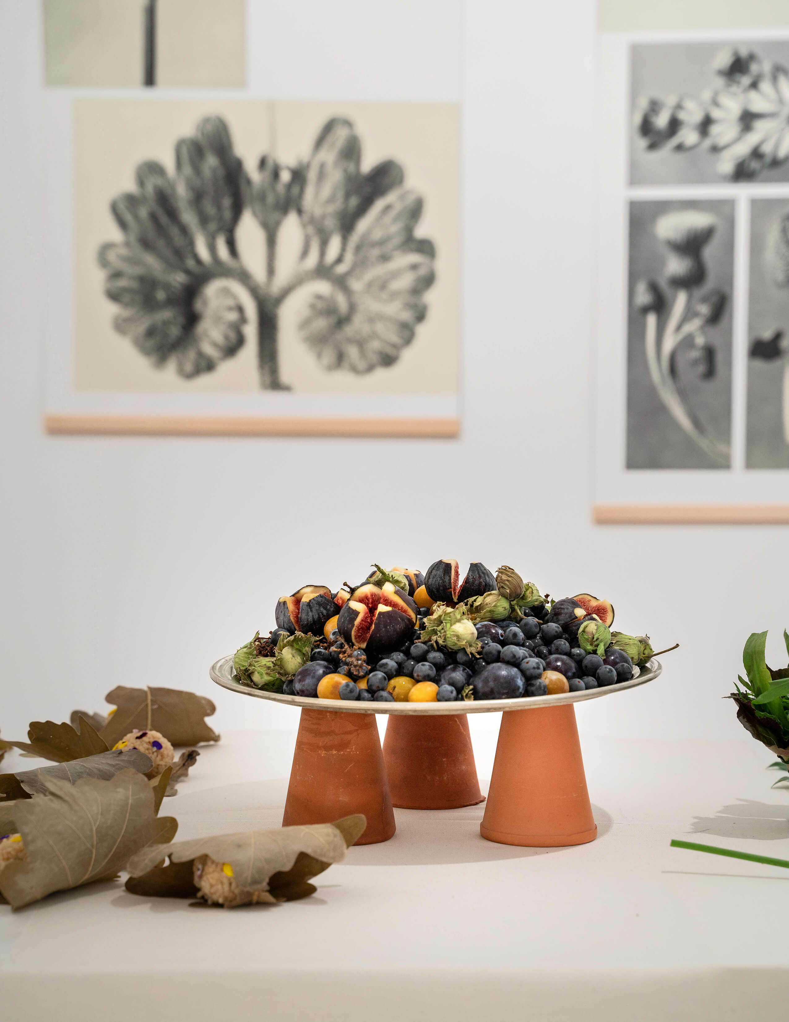 A plate of fruits is displayed next to a few bunches of food that were served with leaves and there are some black and white paintings hung on the wall.