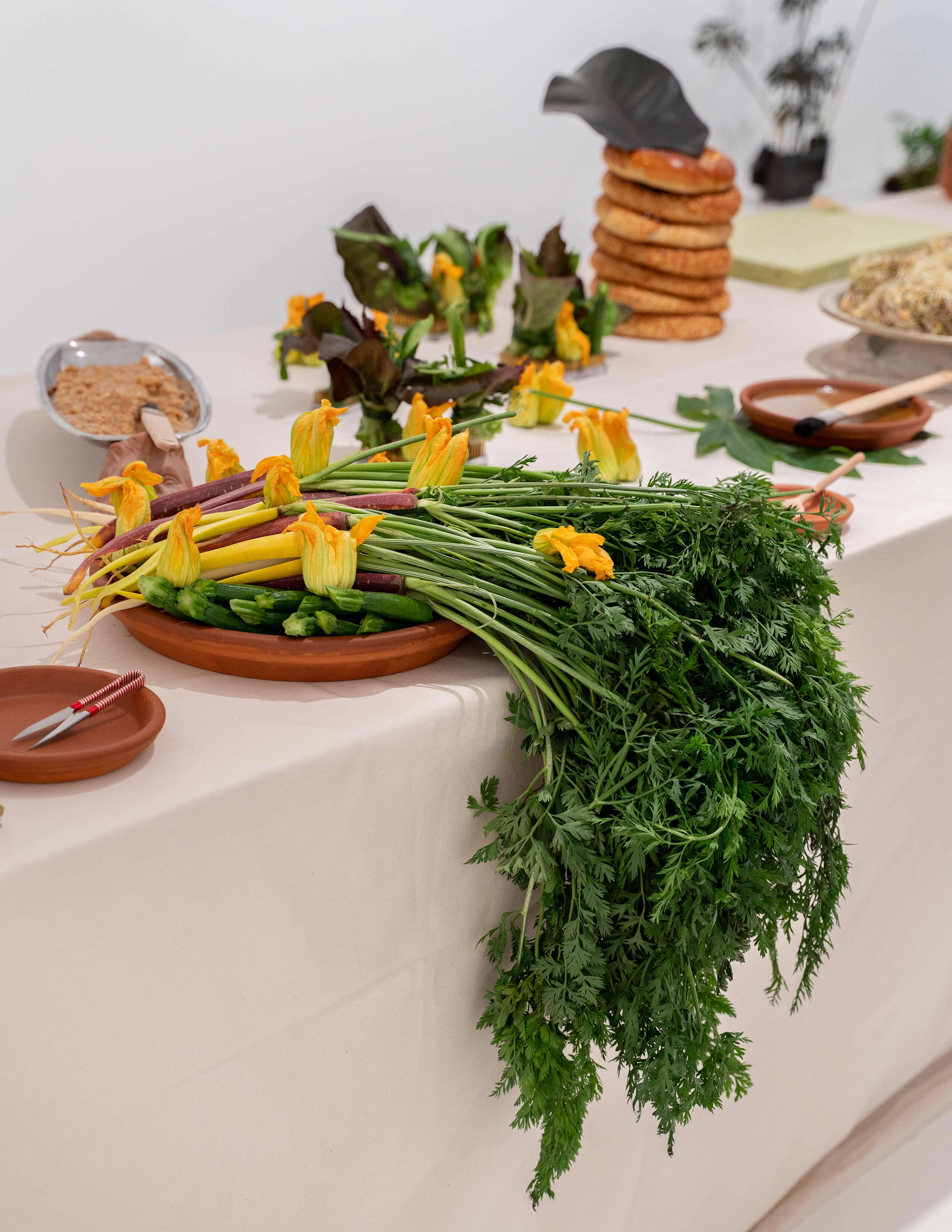 Different kinds of food on the table and there are a bunch of carrots with stems on them, squash blossoms and zucchinis on the plate next to a pair of sewing thread scissors.