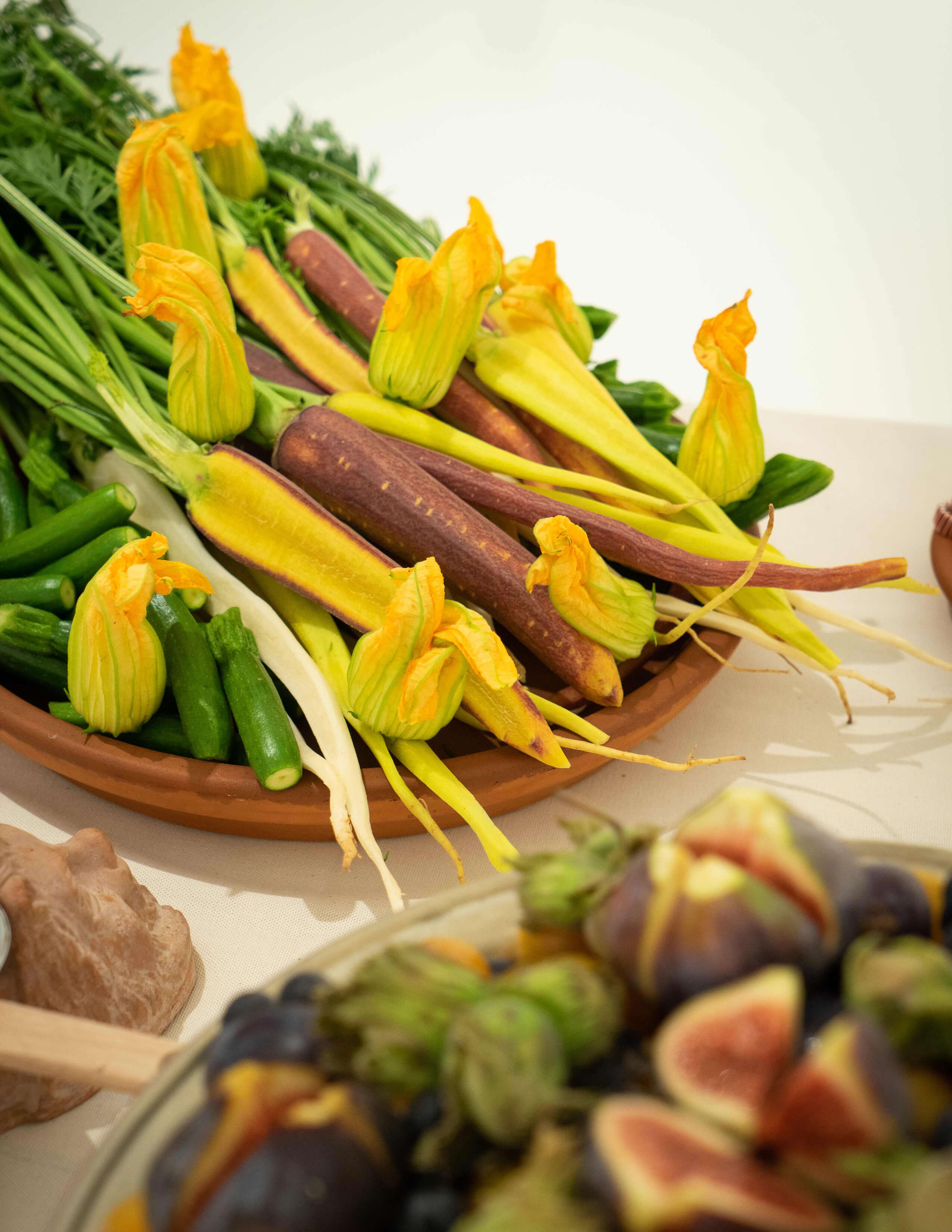 A plate of carrots with stems on it, squash blossoms and zucchinis. Below in the corner, there are figs and other fruits.