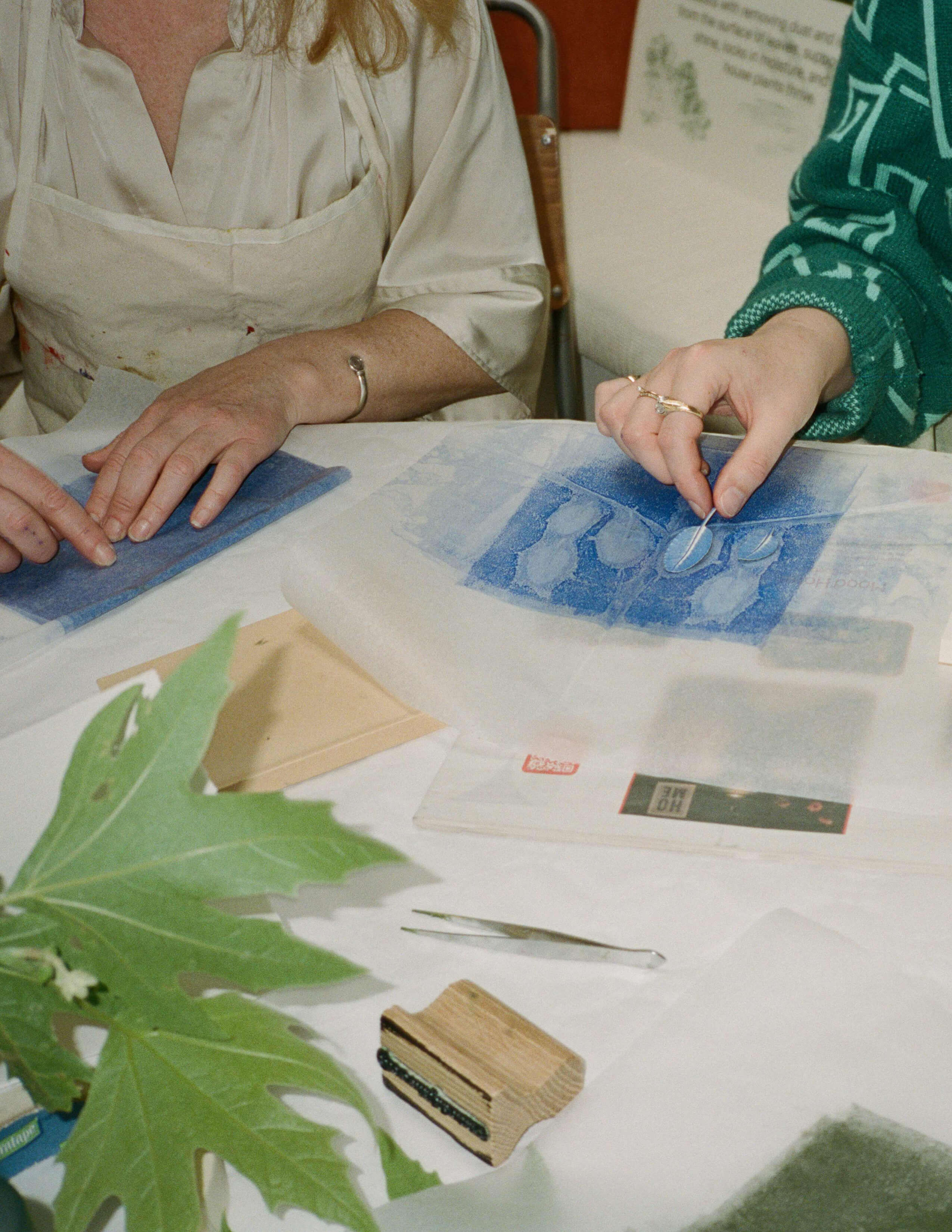 Pia Östlund and another person doing nature printing with a transparent sheet with a blue one on the table. And there is a big piece of papaya leaf, a tweezer and some other tools.