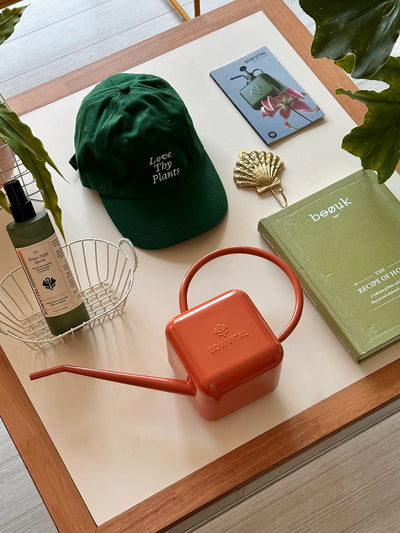 A table has a collection of items neatly arranged on it including a gardener's cap, a terracotta coloured 1.25L stainless steel watering can, and some books.