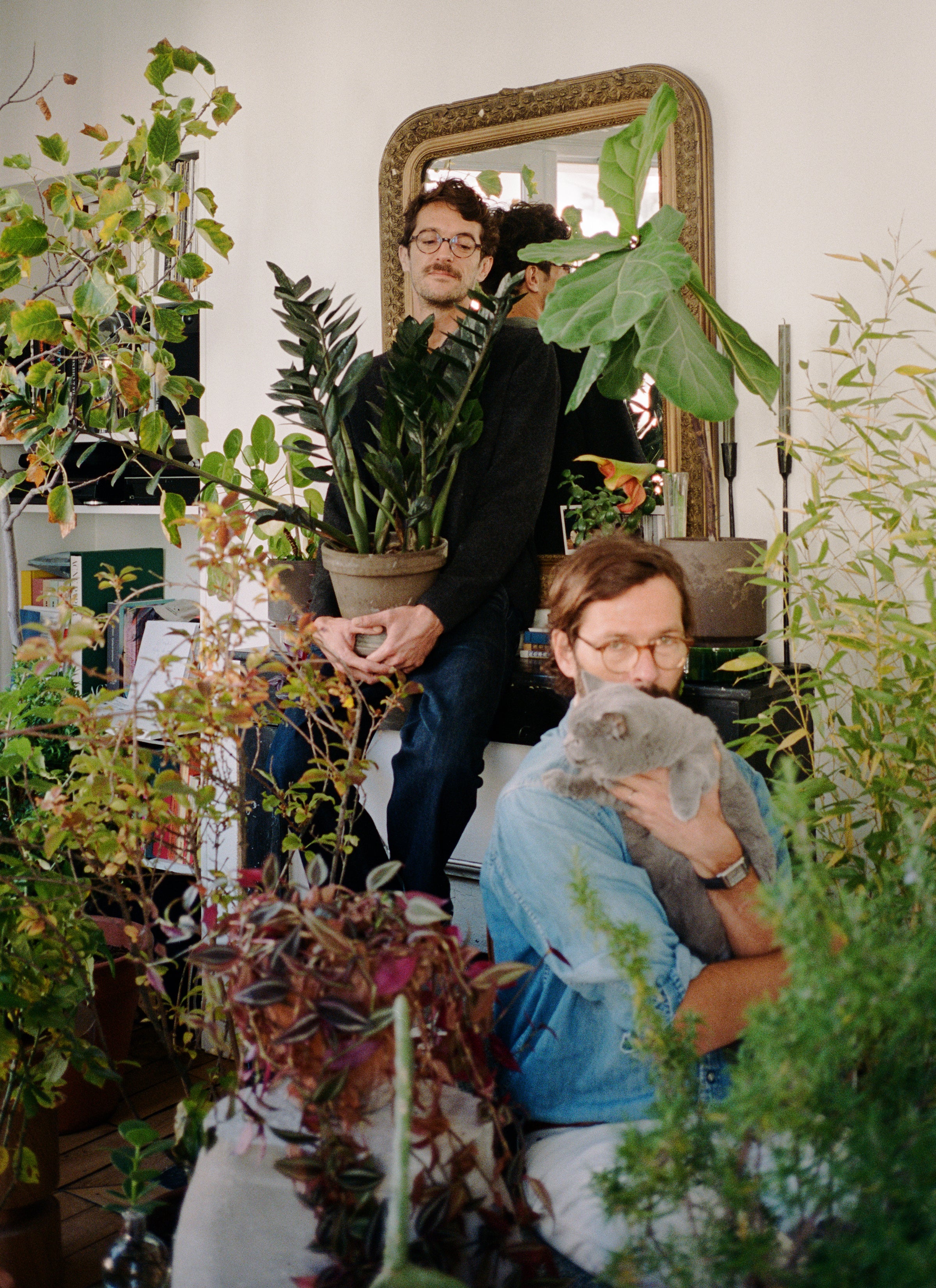 A photograph of Mathias holding their blue-grey cat while Geoffery holding a pot of plants with a collection of house plants surrounded in the room.