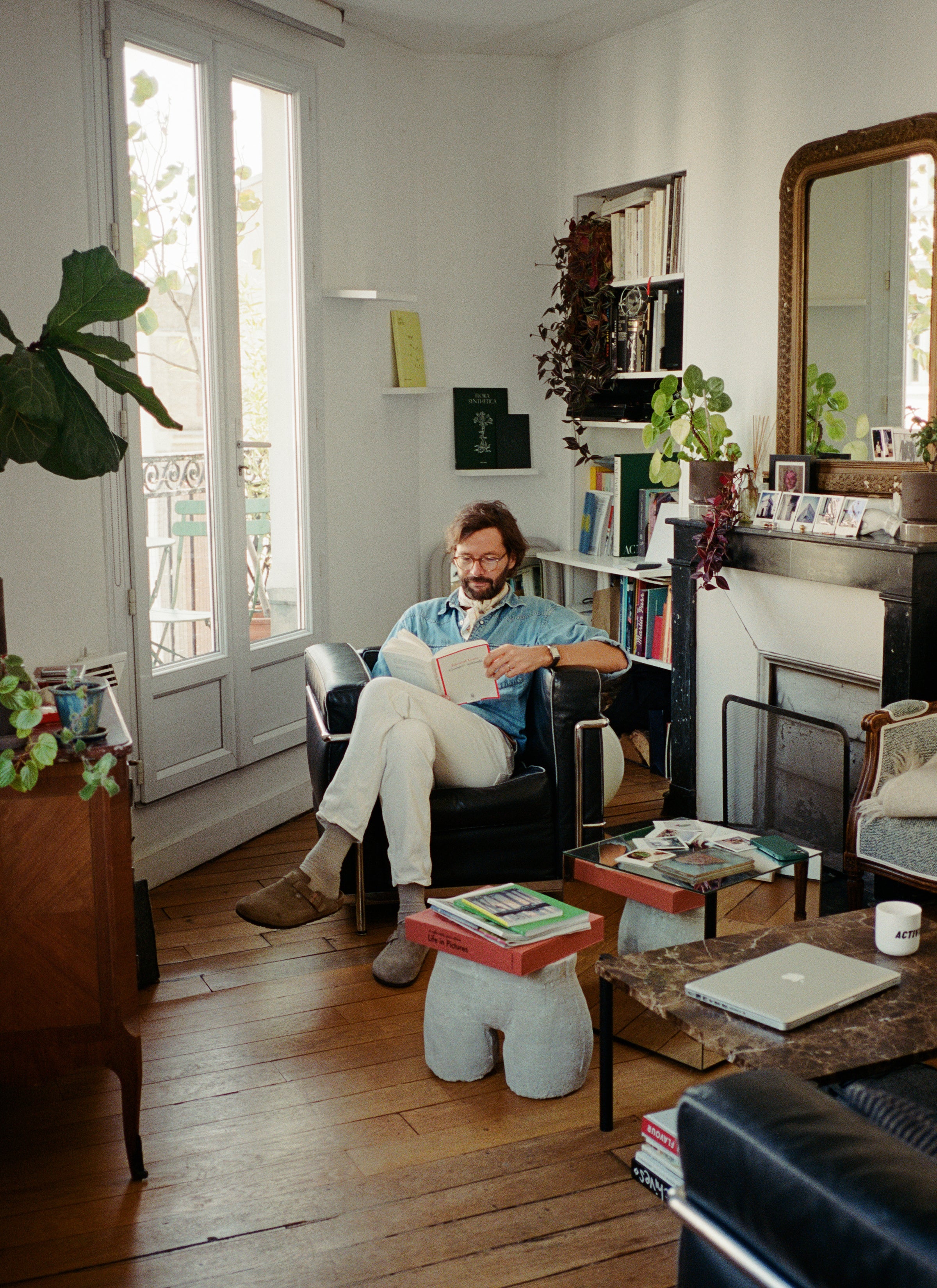 Mathias sat on the sofa reading in the living room which are filled with a lot of books, plants, pieces of furniture and other decorations.