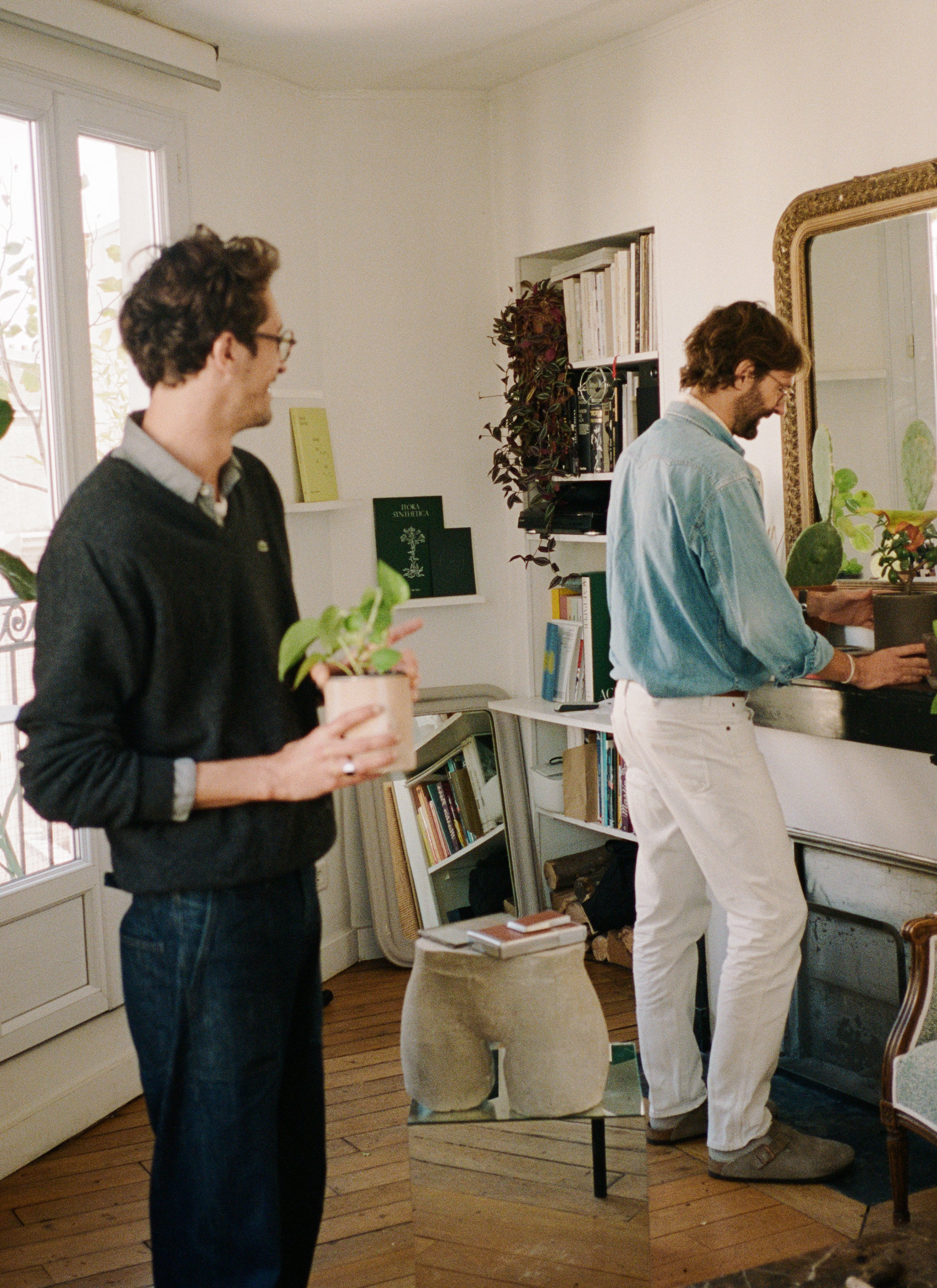 Mathias and Geoffery move pots of plants in their living room with a shelf of books and a buttock-like sculpture with books above it.