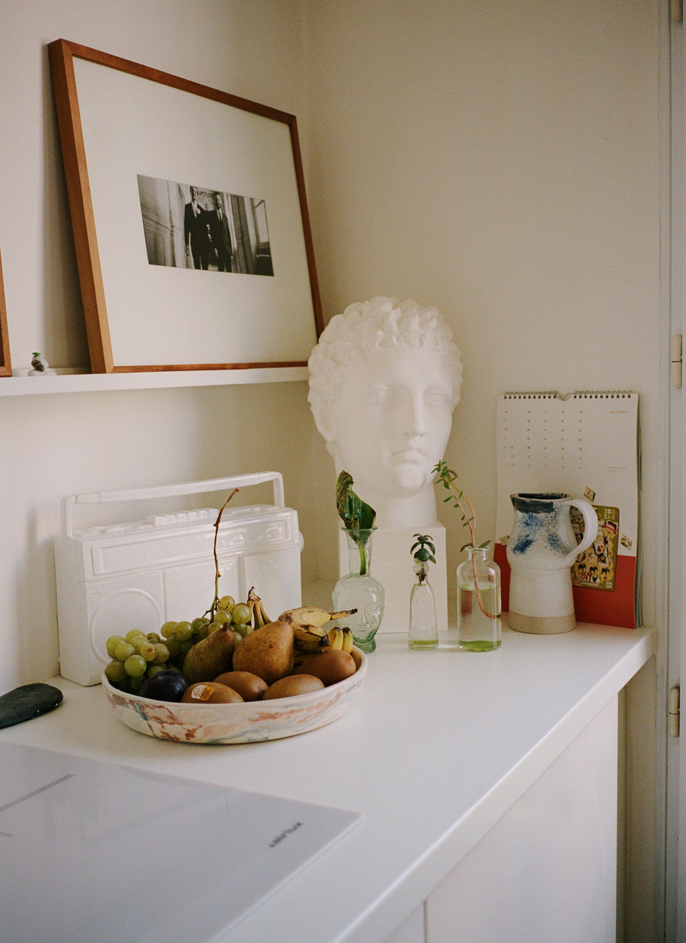 On the table, there is a bowl of fruits with bananas, grapes, plums, pears and kiwis, a white radio, a head statue, three plants growing in different shapes of glass vases, a ceramic-like jug and a calendar. Also a framed black and white picture on the wall shelf.