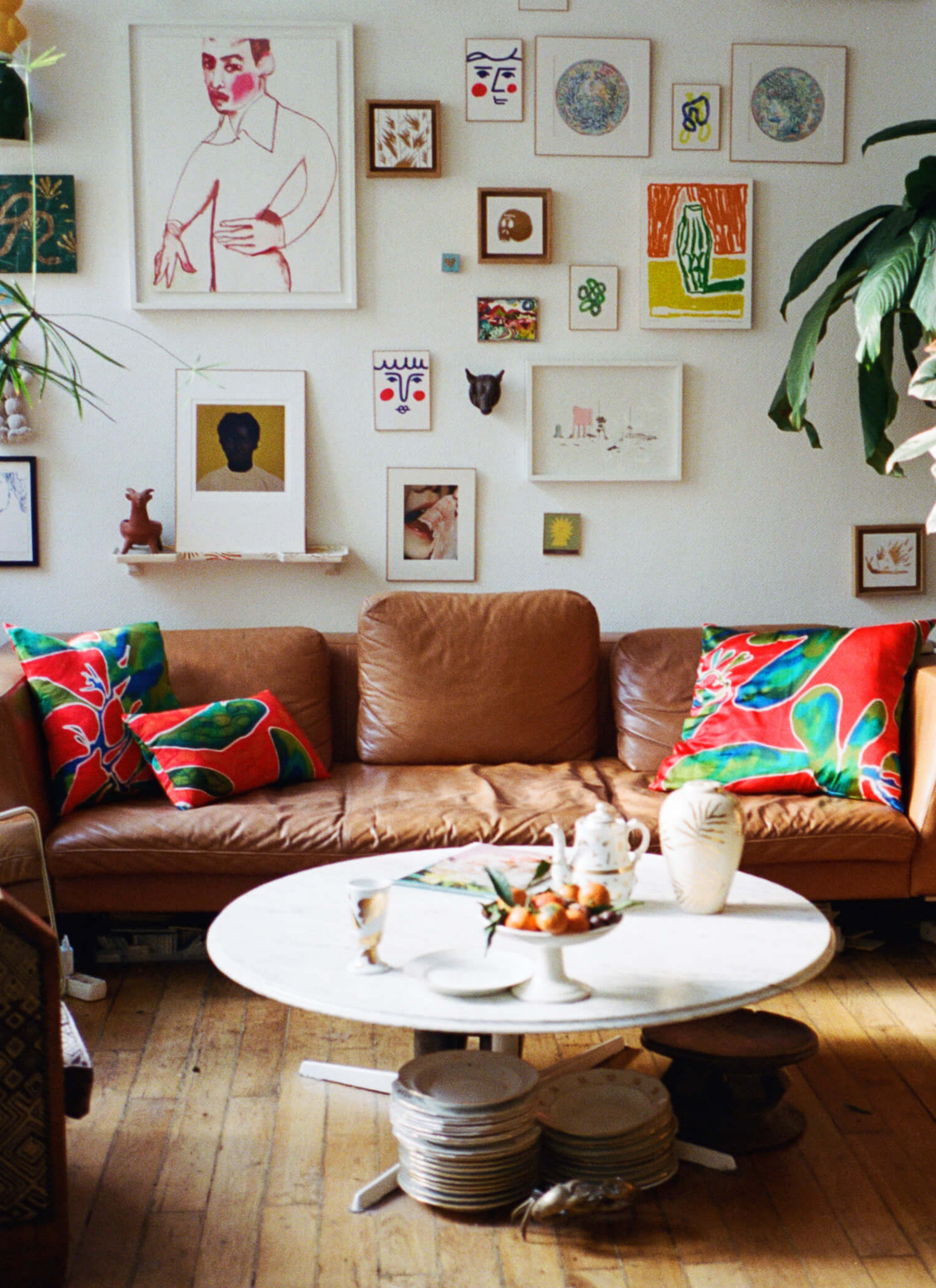 A sofa with some vibrant colour cushions and a white round marble table with some  plates, a teat pot, a cup, a vase and a book on it. And there are many paintings hung on the wall, piles of plates sit on the wooden floor.