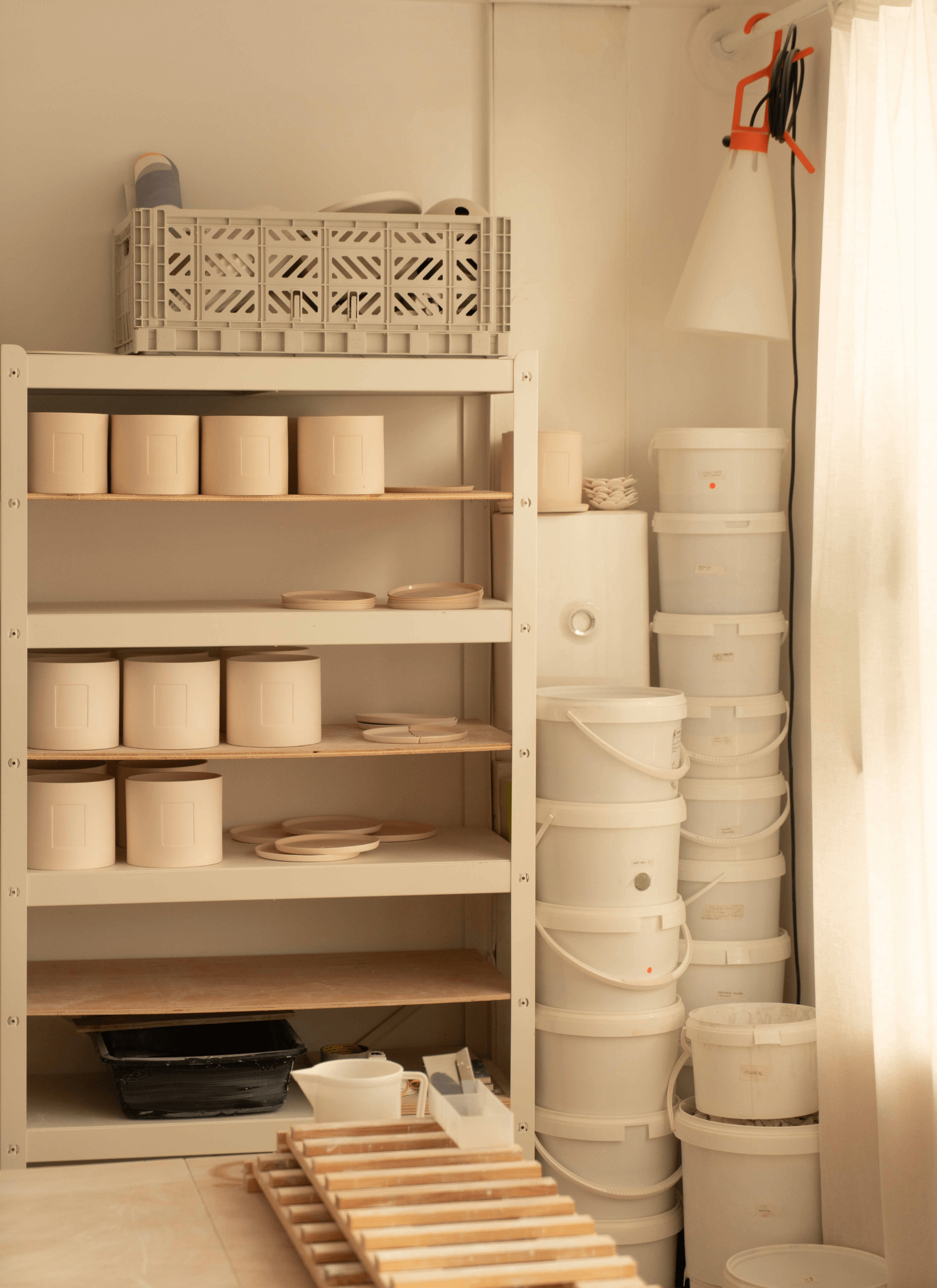 In the corner of the room, stacks of plastic buckets are neatly arranged next to a shelf filled with ceramic pots and saucers. Wooden crates are scattered on the floor nearby, adding to the rustic charm of the space.