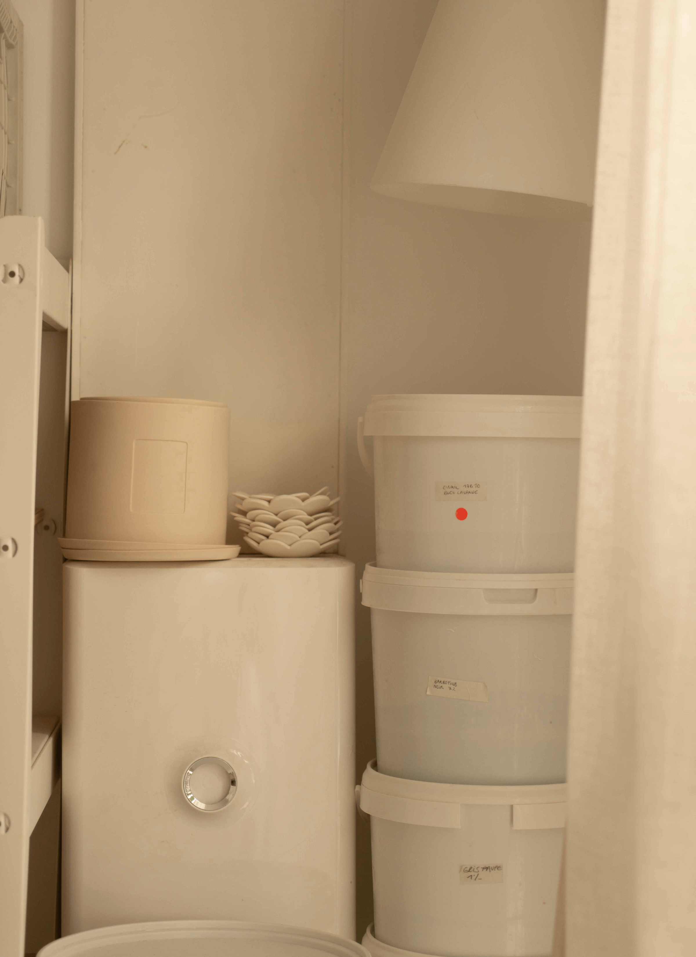 Against the wall, a few plastic buckets are stacked neatly beside a white box. Resting atop the box is a ceramic pot accompanied by ceramic saucers and stacked germination discs. The arrangement showcases a blend of materials, from plastic to ceramic, suggesting a workspace where practicality meets creativity.