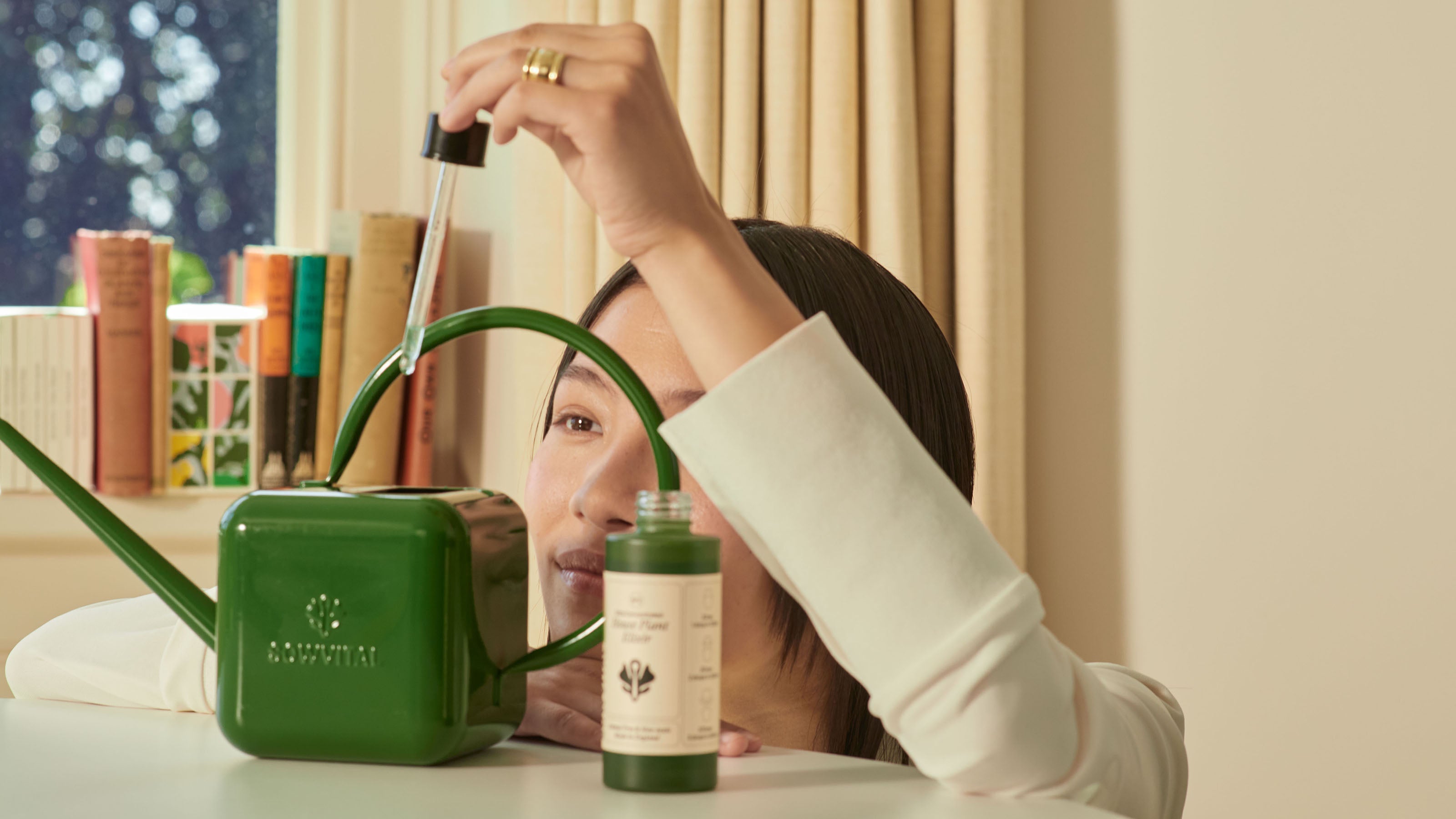 A female model looks closely at the dropper of the House Plant Elixir as she drops it into a Sowvital watering can. She is in a well lit but cosy looking room. The watering can is dark green stainless steel and stamped with the Sowvital logo.