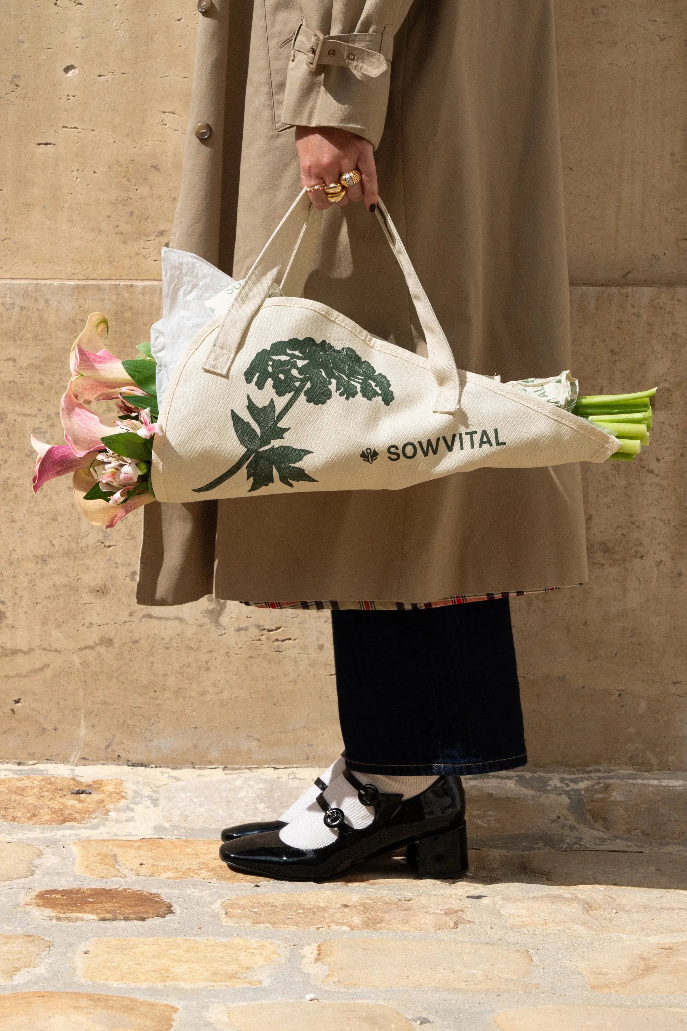 A woman stands in a long beige coat wearing shiny black shoes and white socks. She is holding a cotton tote bag specifically designed for carrying bouquets of flowers. There is a large bouquet contained within.