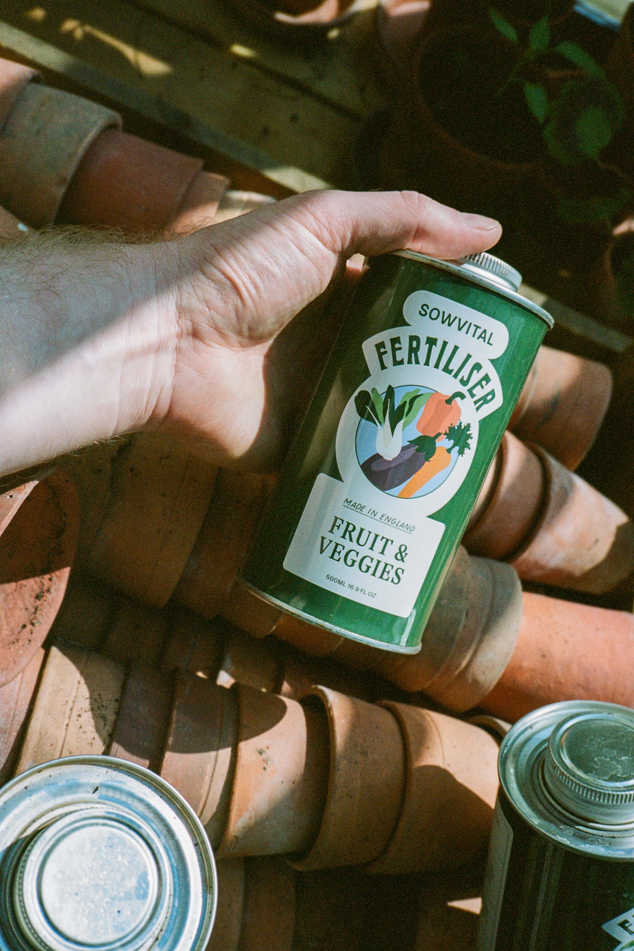 A hand holds a dark green metal tin of Sowvital's Fruit & Veggies fertiliser above a crate of small, stacked terracotta pots.