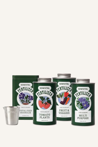 Four different green metal tins of garden fertilisers sit alongside a small metal measuring cup in front of a beige background.