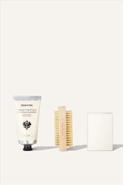 The contents of the Gardener's Hand Care Kit. 3 products sat in a row, The Gardener's Hand Cream, a wooden nail brush with "Love Thy Plants" engraved into the side, and a white bar of soap. The background is beige.