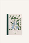 An A6 notebook with a beige cover and green cloth spine. The cover shows a green and blue illustration of a collection of glass flasks, all with different propagations and plant cuttings in them. The background of the image is beige.