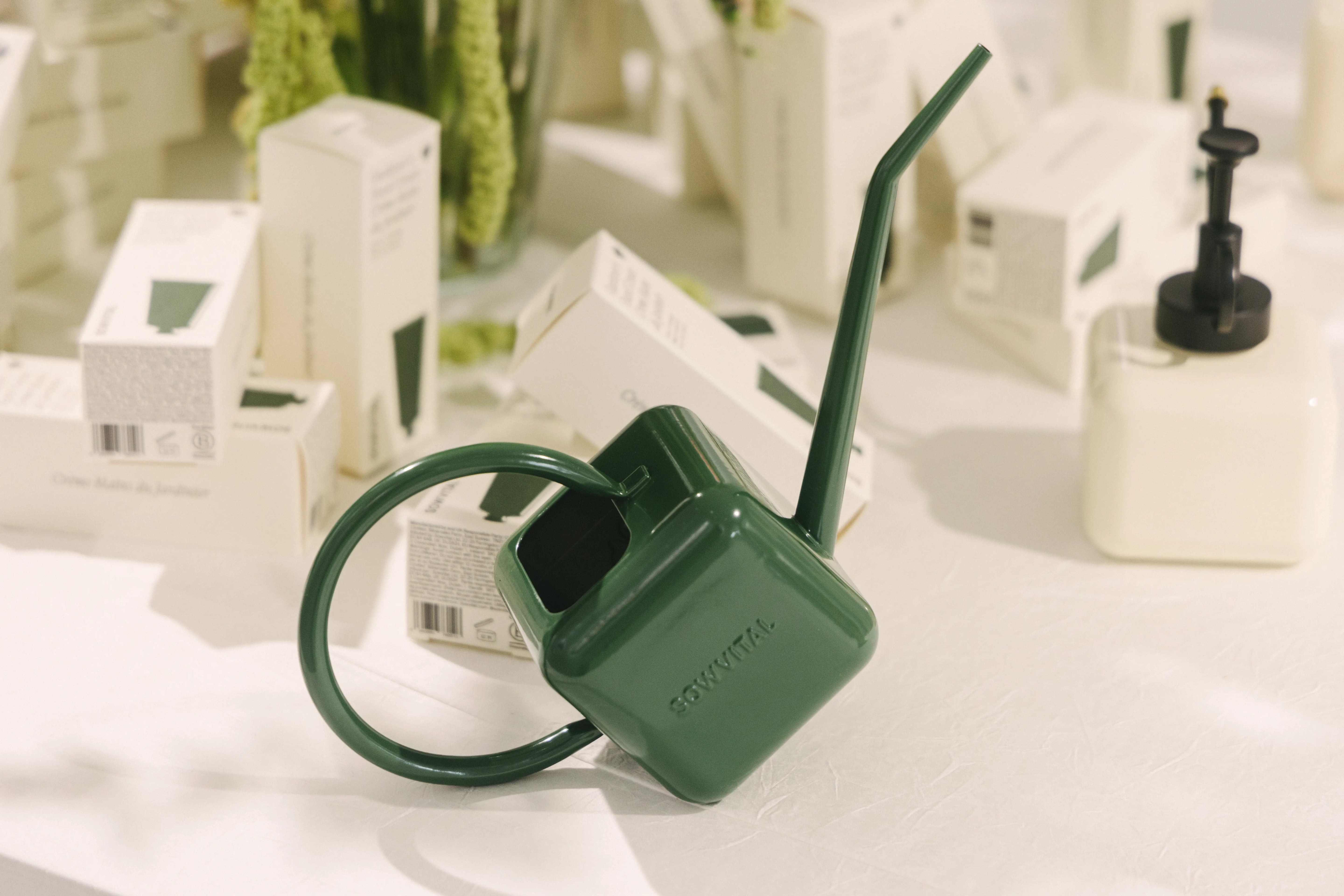 A stainless steel green watering can is seen resting on its handle, the background is filled with boxes of hand cream, and a beige mister is seen to one side.