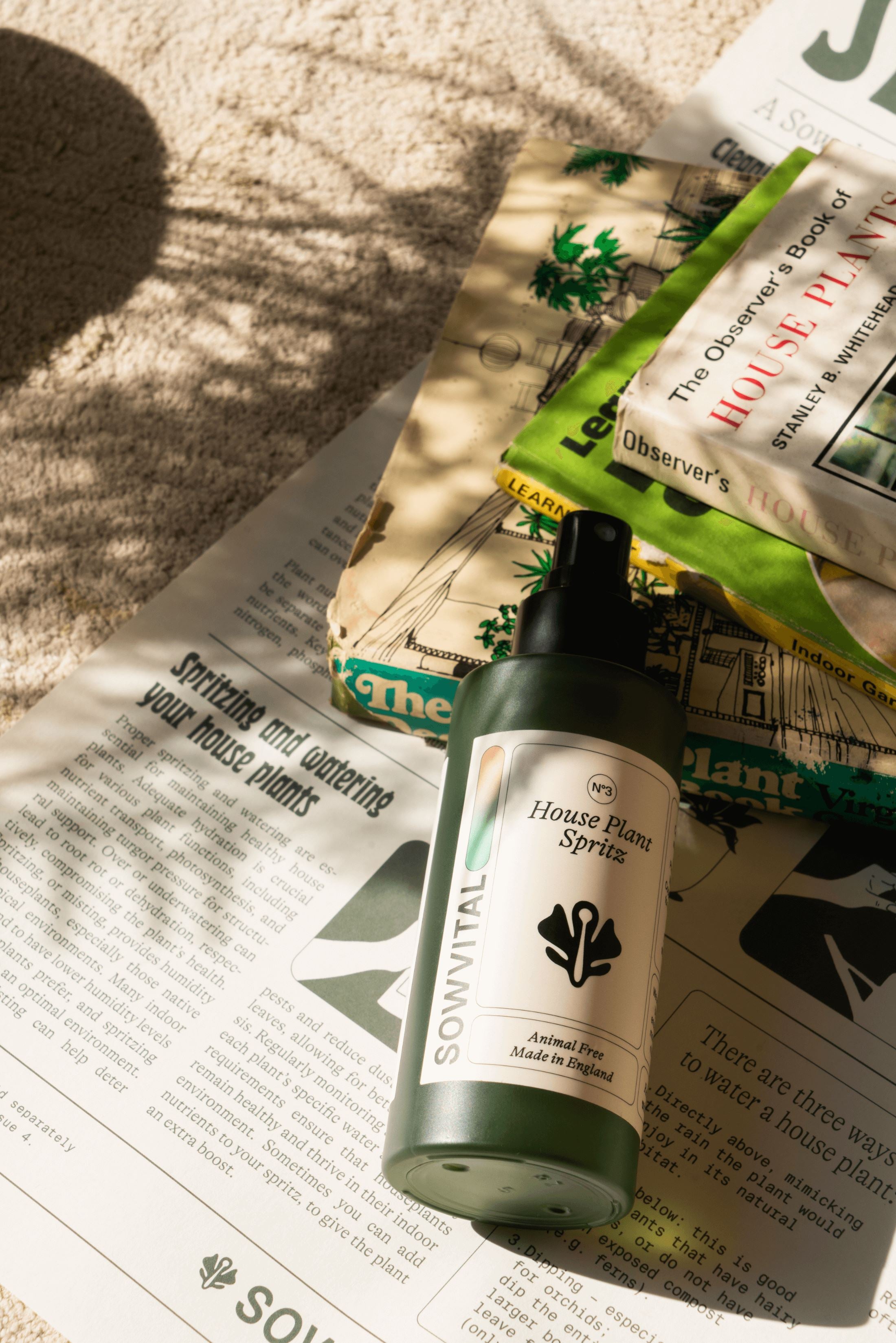 The House Plant Spritz, a 100ml green bottle with a beige label and black spray top lid, sits in dappled sunlight on a beige carpet, surrounded by plant care books and posters.