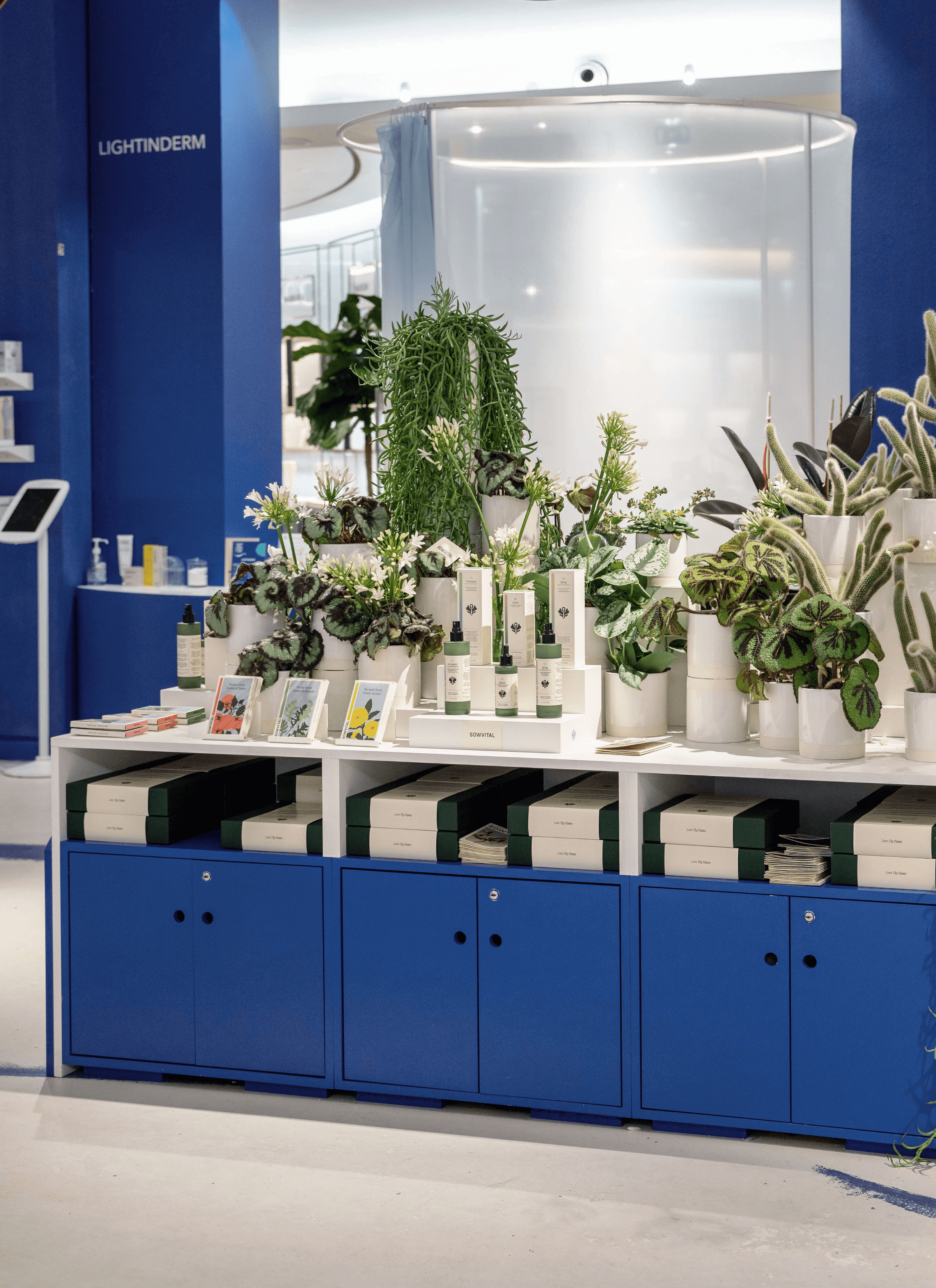 A display area in Le Bon Marché showcases Sowvital's products arranged neatly on the surface. Behind the products, a variety of potted plants, flowers, and cacti are used to create a lush and colorful backdrop. The bottom section features blue-painted cupboards, serving as a contrasting base for the display. Inside the cupboards, many three-step gift sets are neatly stocked, adding to the visual appeal and accessibility of the products. 