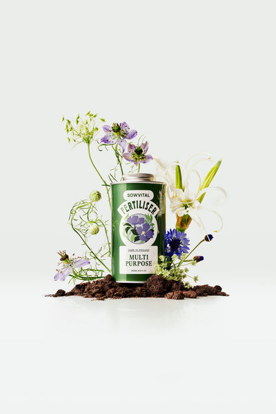 A green metal tin of Sowvital's Multi Purpose fertiliser, sat atop a small mound of soil and surrounded by flowers and plant material.