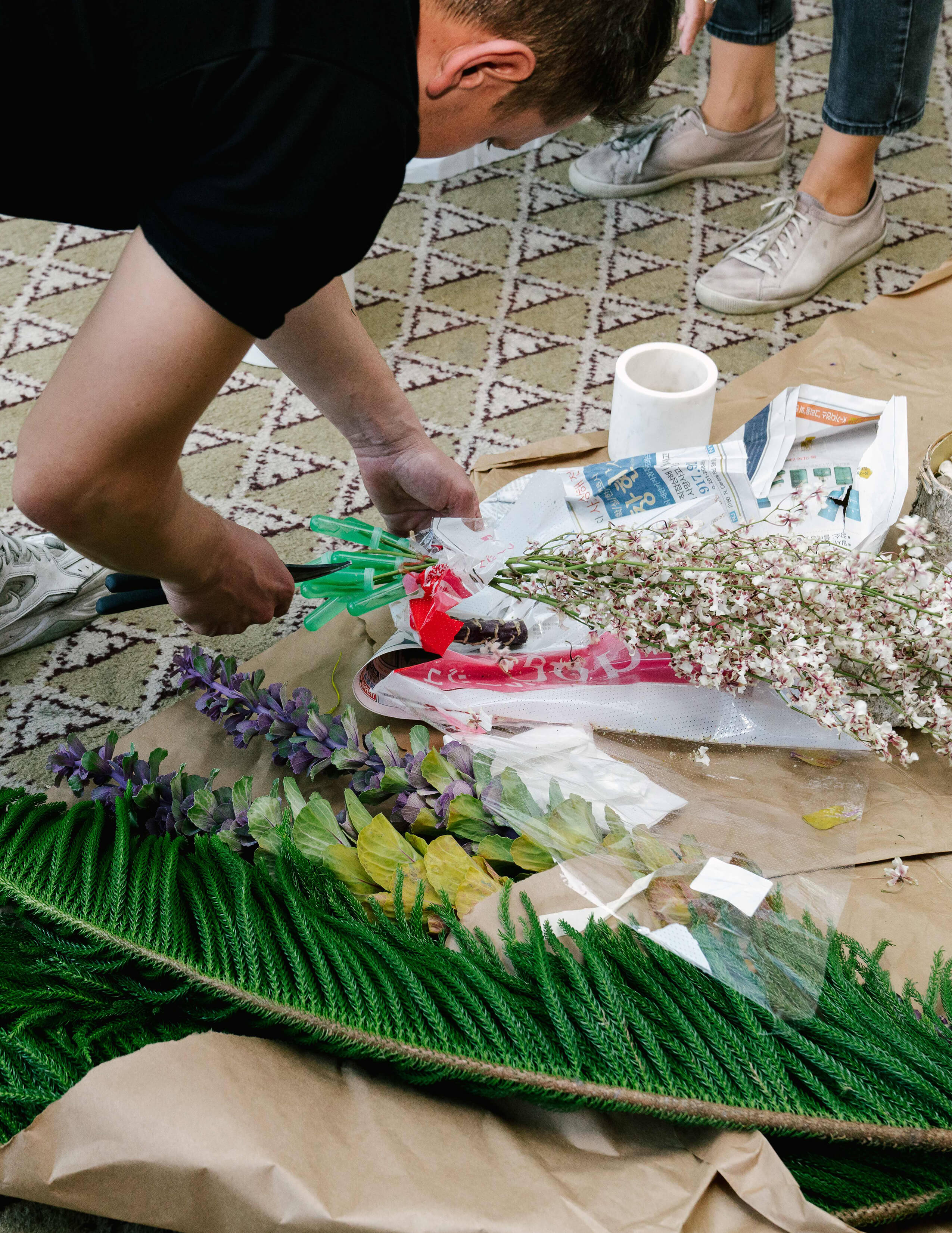 A person bends down his waist with secateurs in his hand and approaches a bundle of flowers on the floor while there are other kinds of plants and flowers lying around.