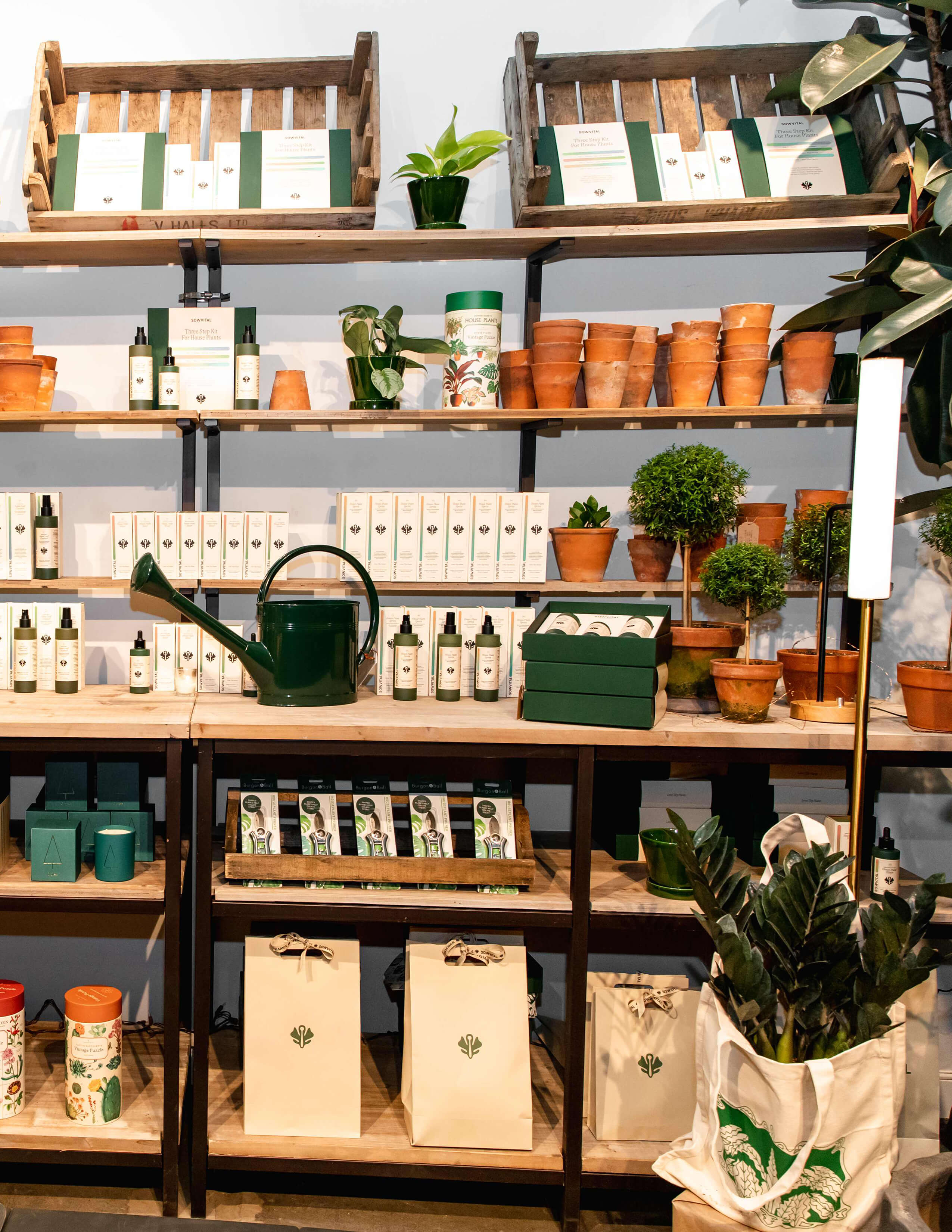 A collection of Sowvital house plant products is presented nicely on the shelf surrounded by some potted plants and there is a green water can in between the products too. Some gifting bags are on the lower shelf.