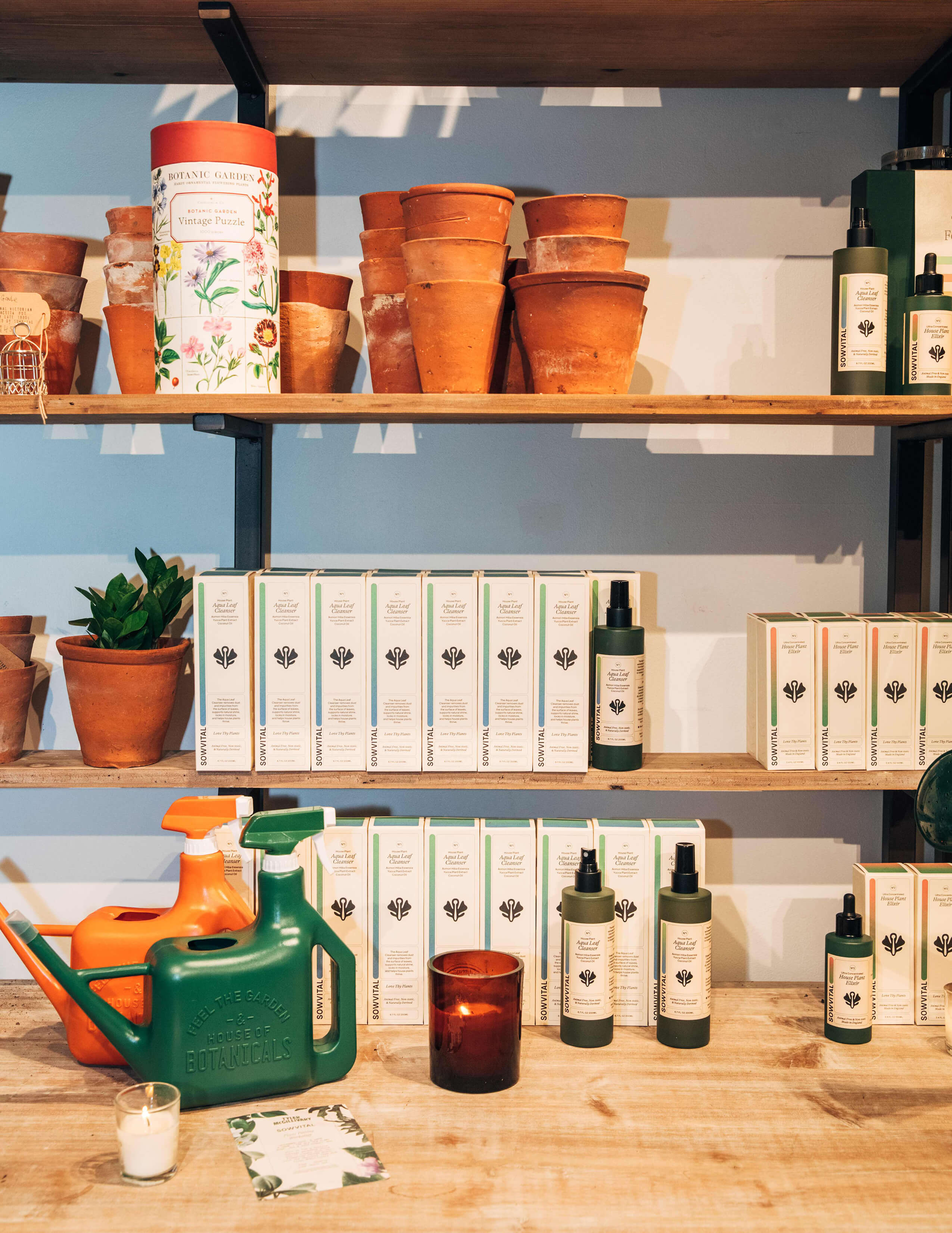A shelf of Sowvital house plant products and terracotta pots while there are orange and green water cans, and two lightened-up candles around on the surface.
