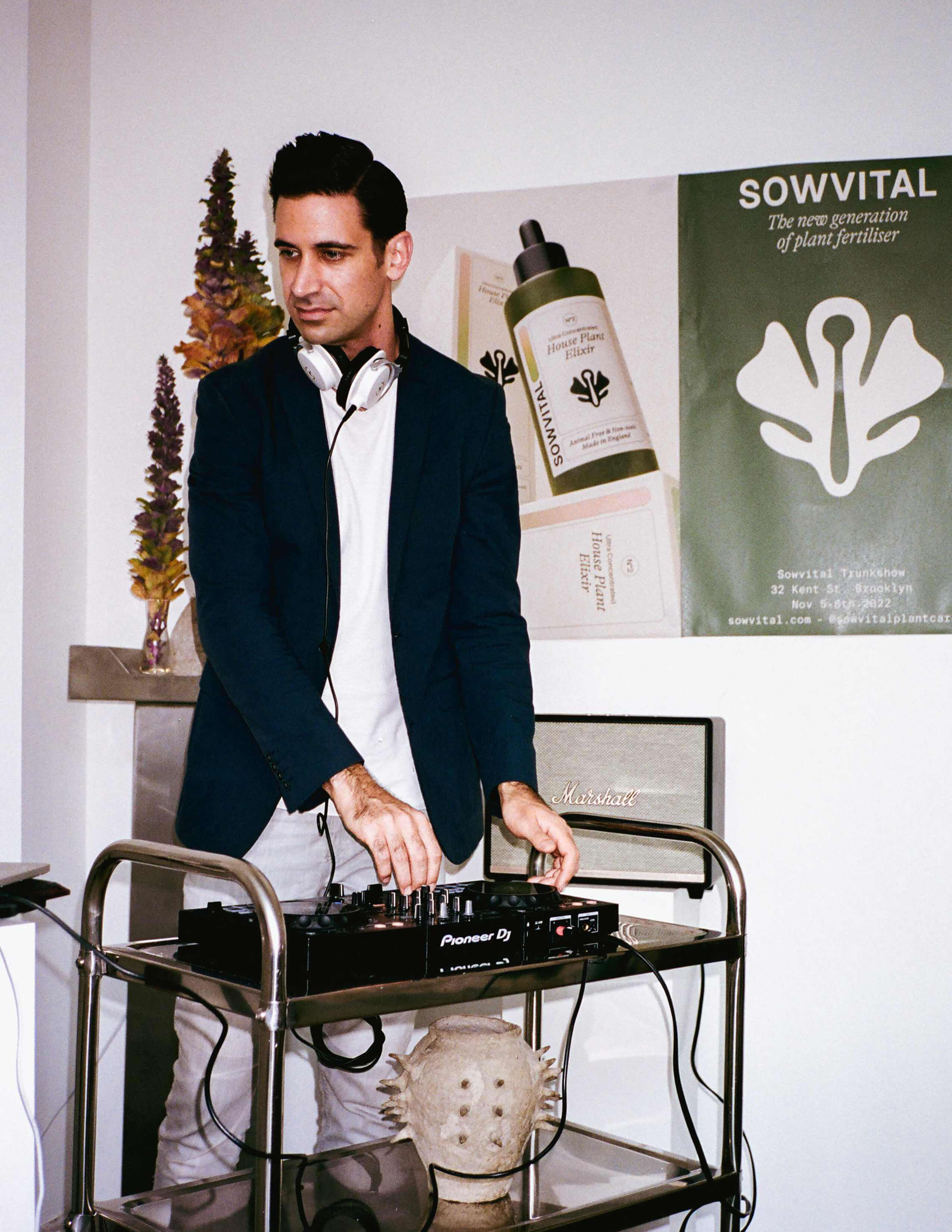The DJ in a black blazer is djing on his Pioneer deck while there are Sowvital product posters and some plants behind him.