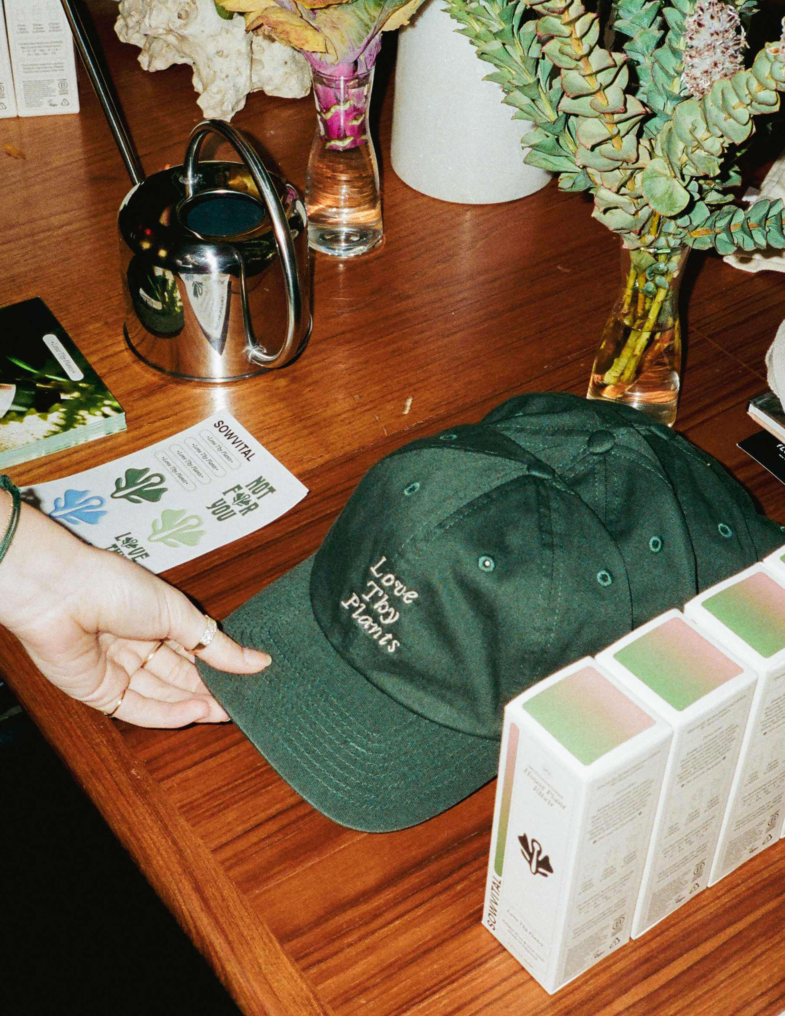 Different kinds of plants were inside separated glass bottles while there was a silver water can, Sowvital branding stickers, ‘Love Thy Plants’ caps and house plant products on the table.