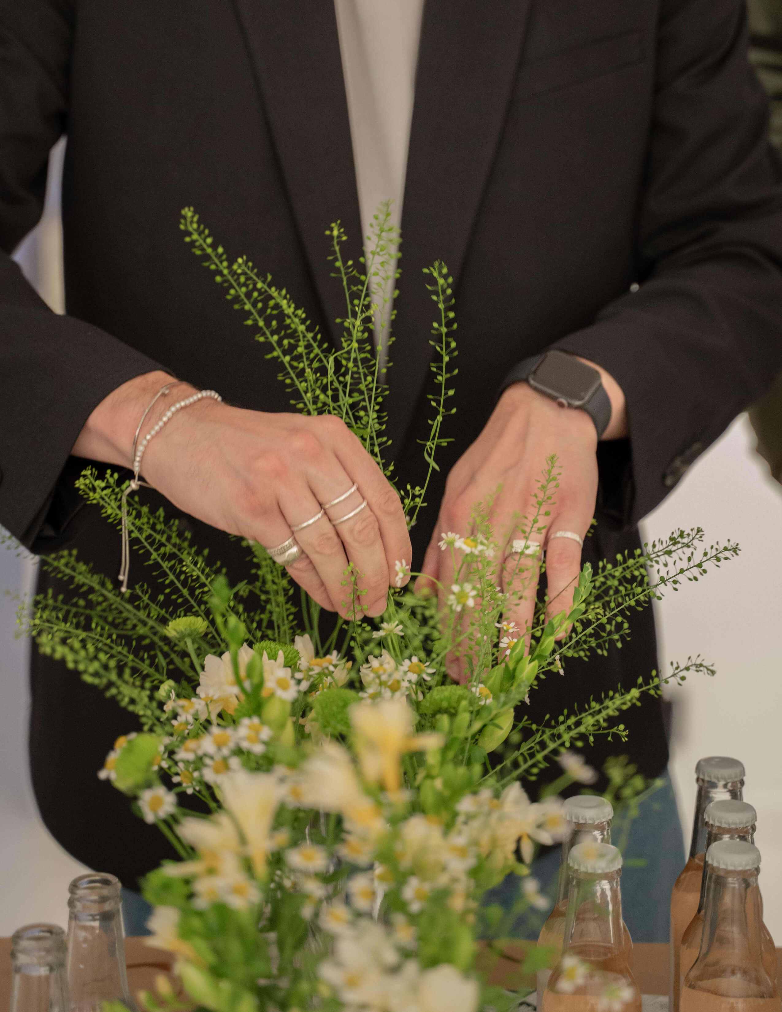 A person with a black blazer organises the plants and flowers next to bottles of alcoholic drinks.