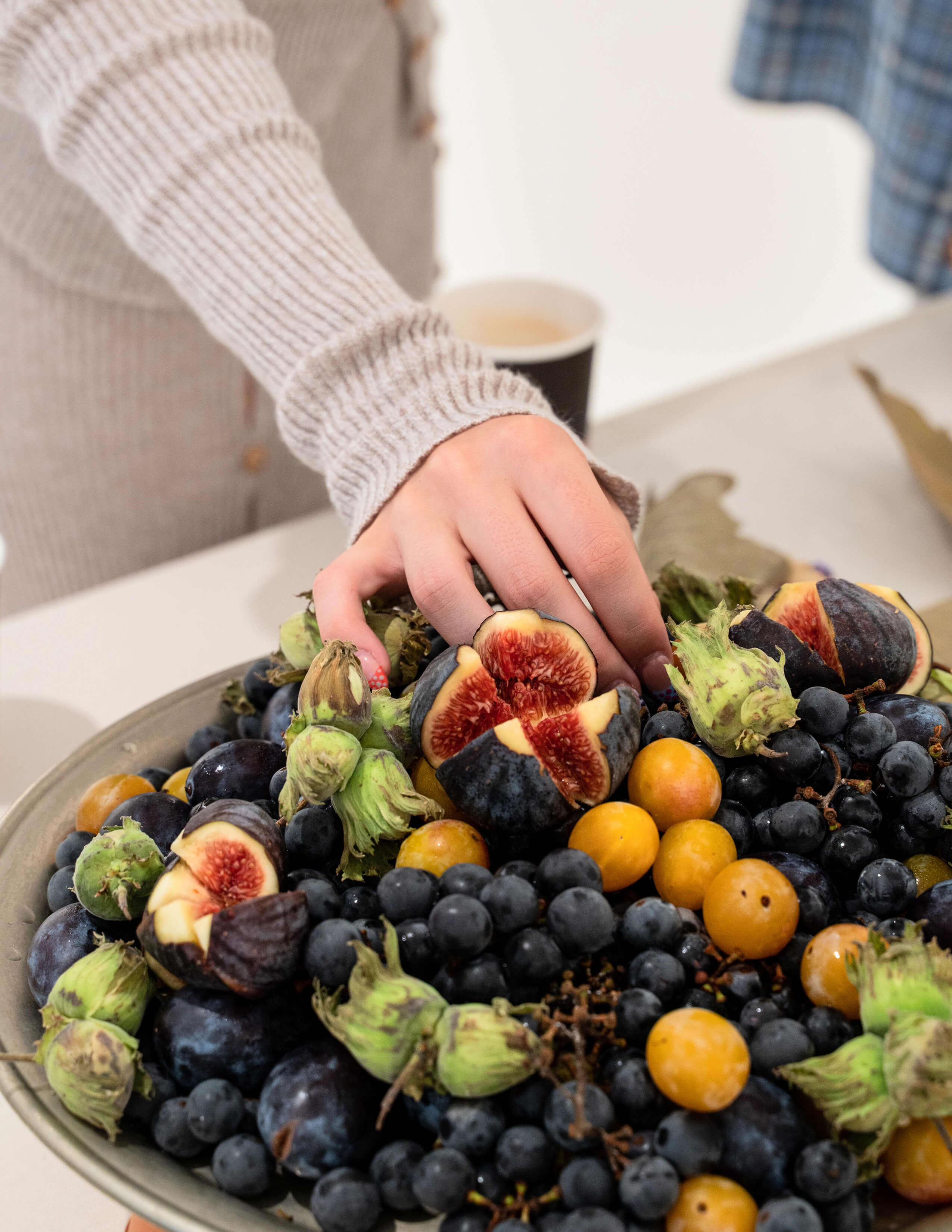 A hand on a plate of fruits filled with blueberries, grapes, figs and flower buds.