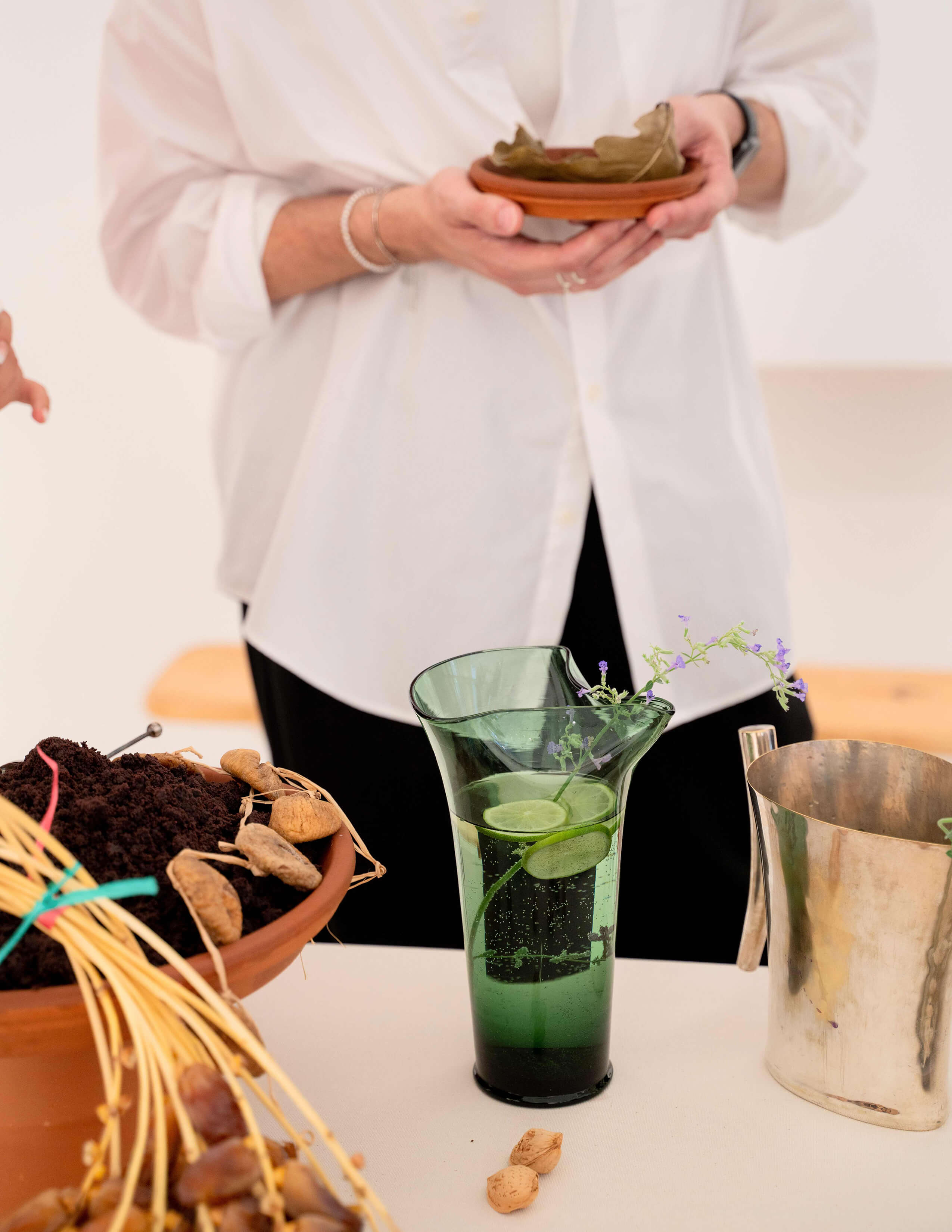 A person stood behind a green glass of drink that contained 3 slices of lemon and a few stems of flowers in it. There are two plant seeds, a bowl of soil with mushrooms and a jug on the table.