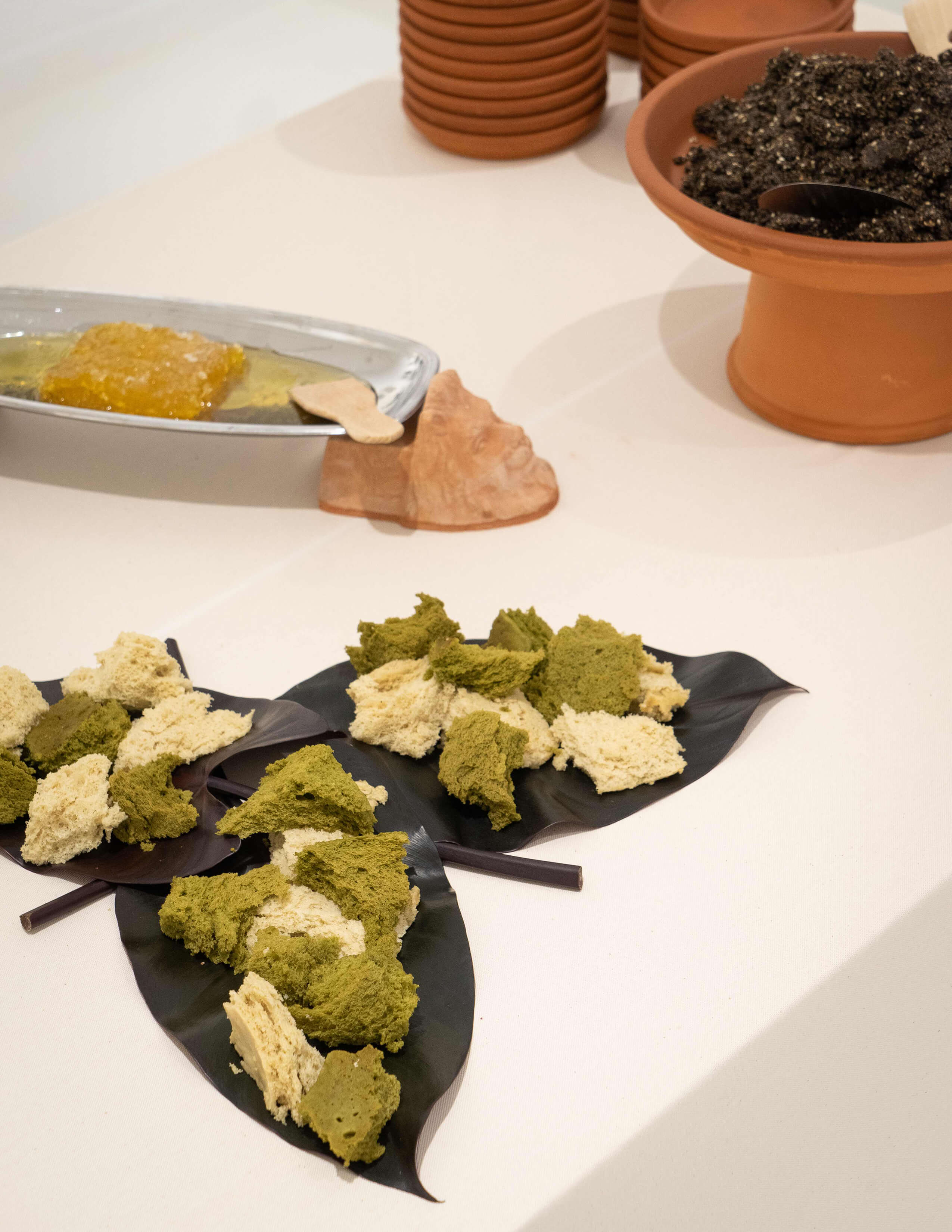 Pieces of ripped bread in green and cream colour served on three dark colour leaves. A bowl of soil and a big chunk of honey-like thing served behind.