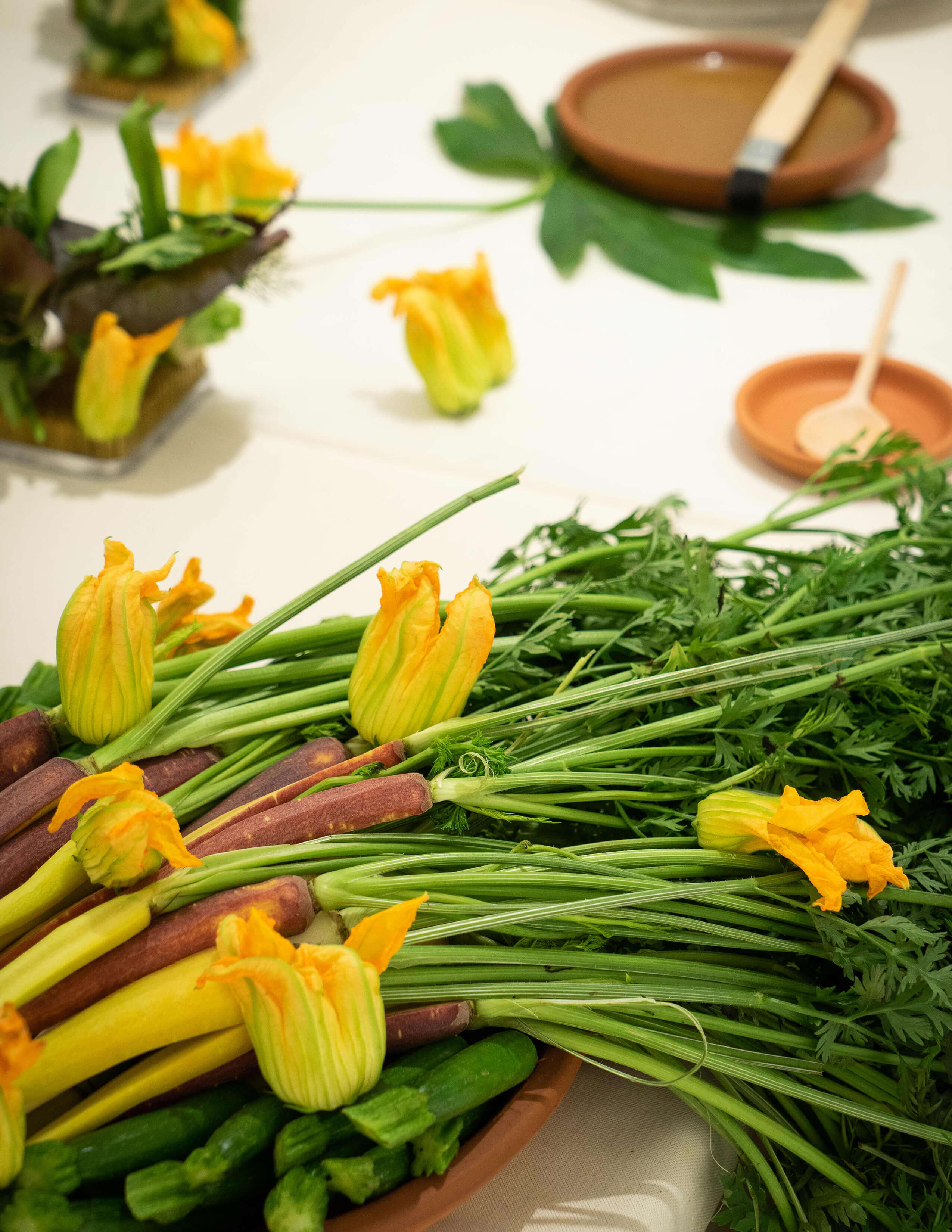 A plate of carrots with stems on, squash blossoms, and zucchinis on the table.