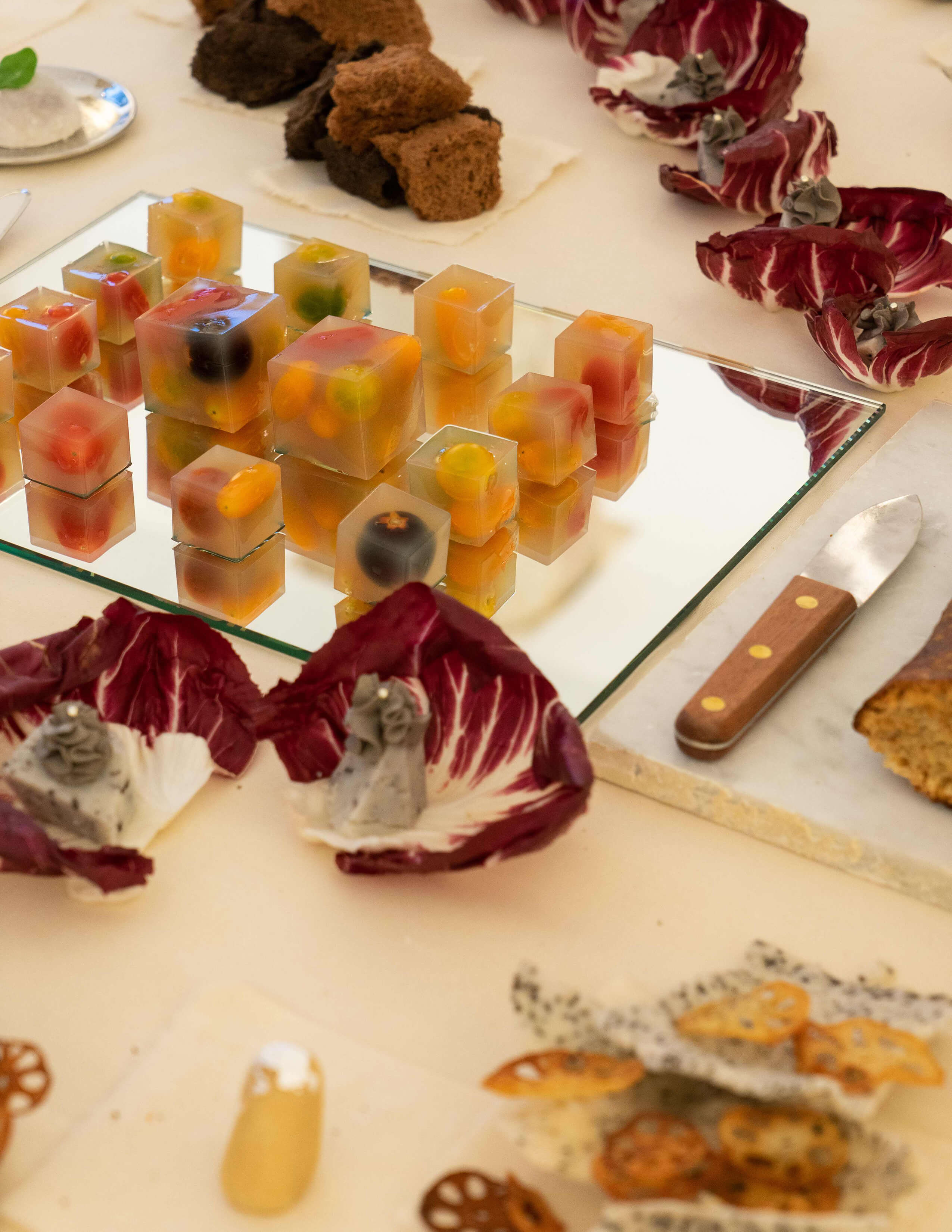 Loads of small jelly cubes filled with different shapes of fruits are served on the glass board next to a bread knife and there are differentfoodsd served on pieces of purple leaves.