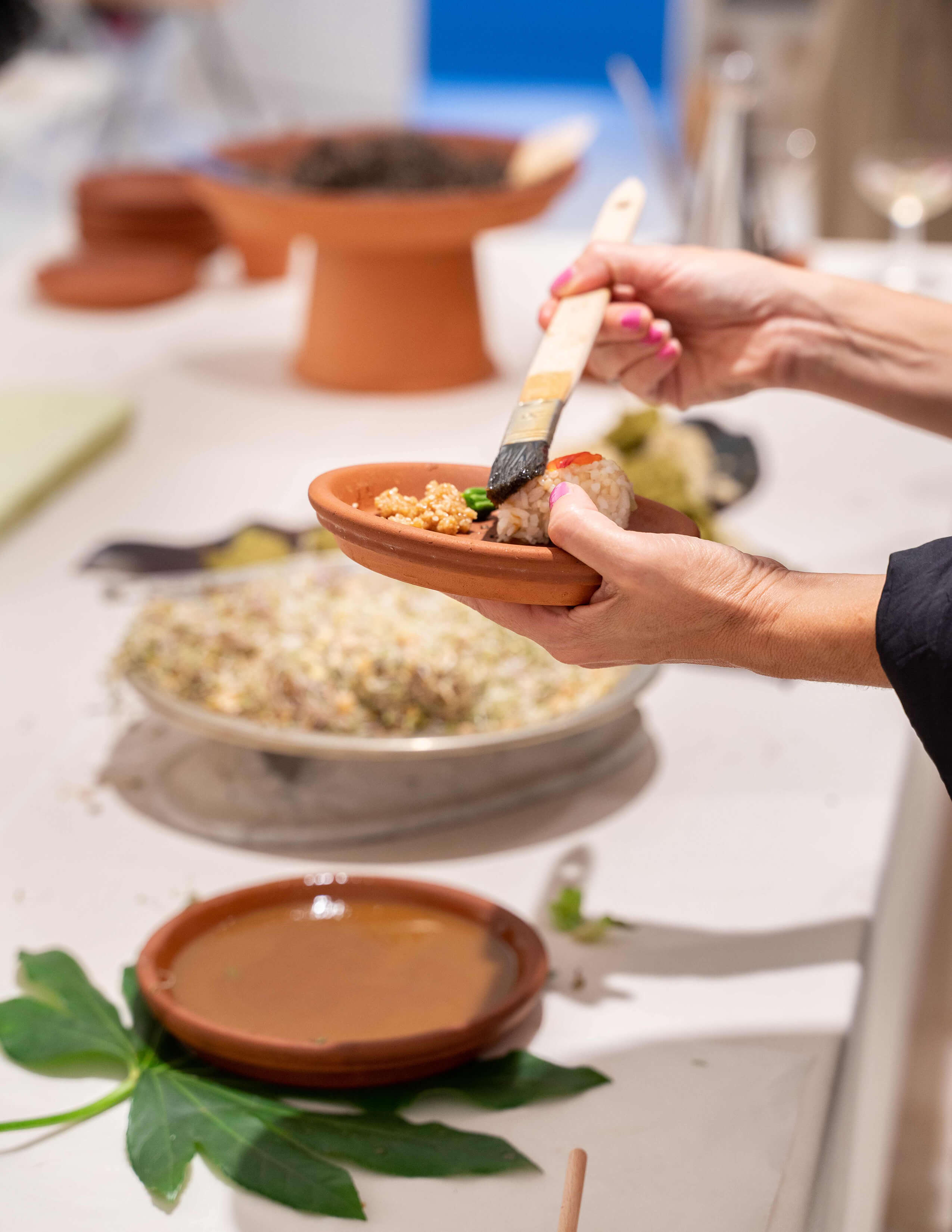 A pair of hands with pink nails, holding a plate of food while brushing sauce on it, and a big piece of leaf is underneath a plate.