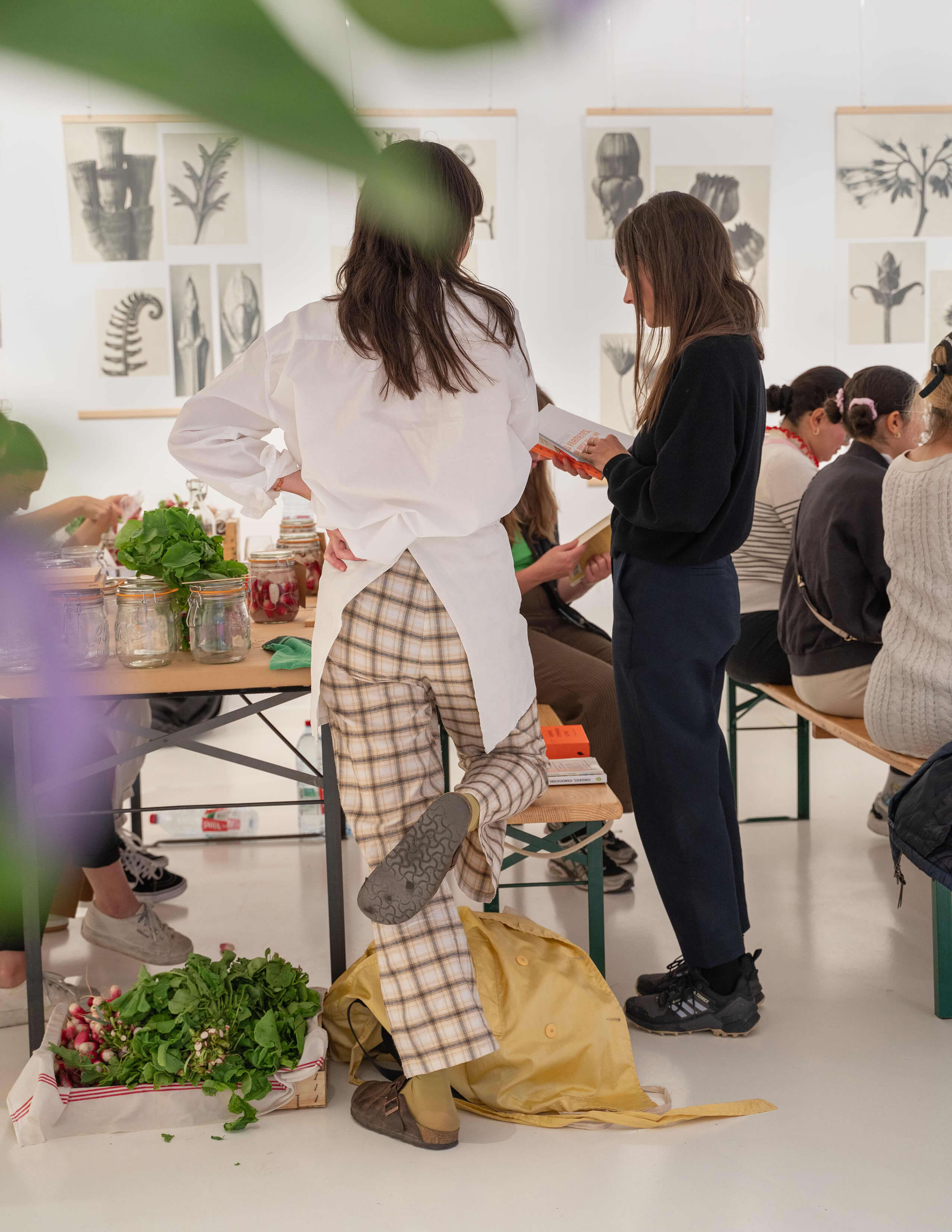 Some people sat down and some stood up in the room. There are some plants, and vegetables on the floor and some are in the jars on the table—a load of pieces of plant theme art behind on the wall.