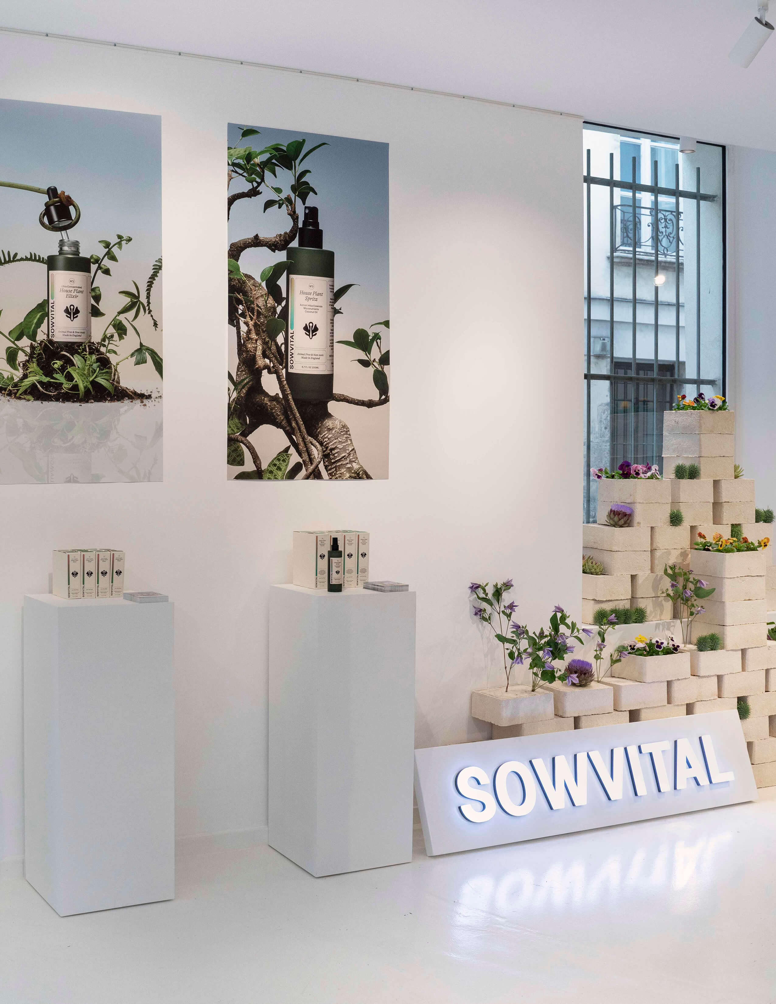 Posters of Sowvital house plant products on the wall and the products on the stands next to the Sowvital logo. Loads of different kinds of plants and flowers on the side with white cubes.