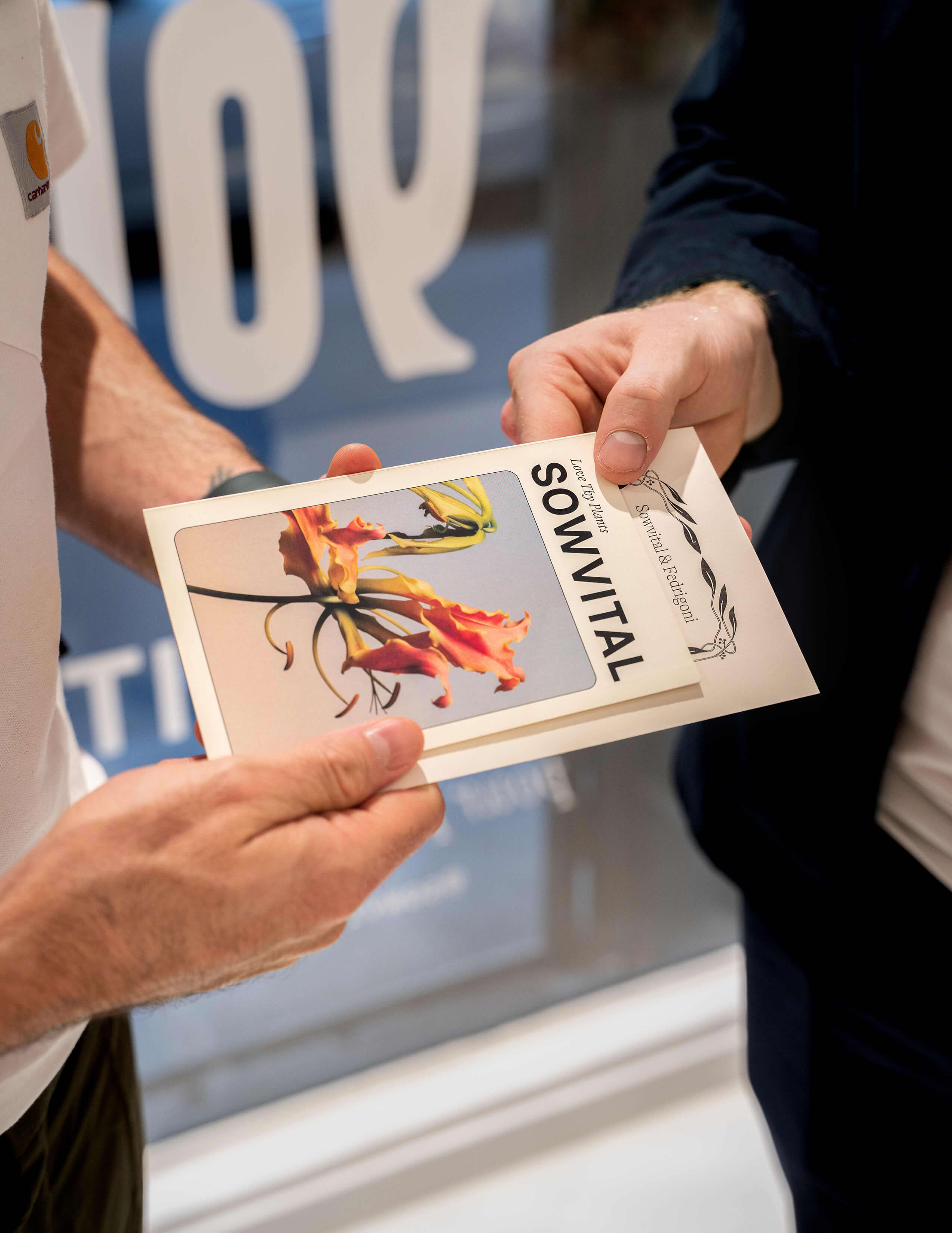 Two pairs of hands holding a card with Sowvital’s logo on it with an orange-pinky flower on the cover.