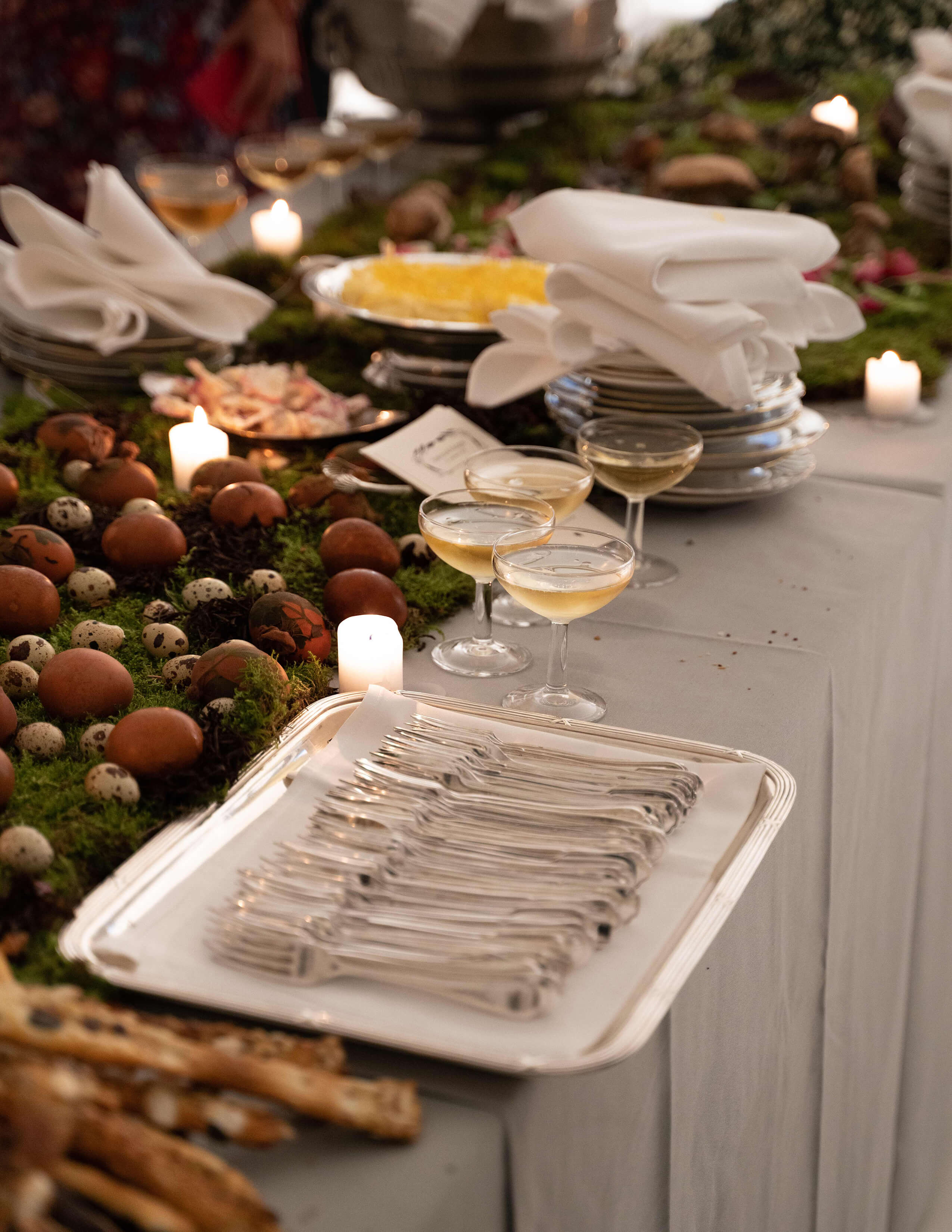Plenty of forks are displayed on top of a white sheet of silverware and there are different kinds of eggs displayed on moss and a couple of glasses of drinks, plates and serviettes.