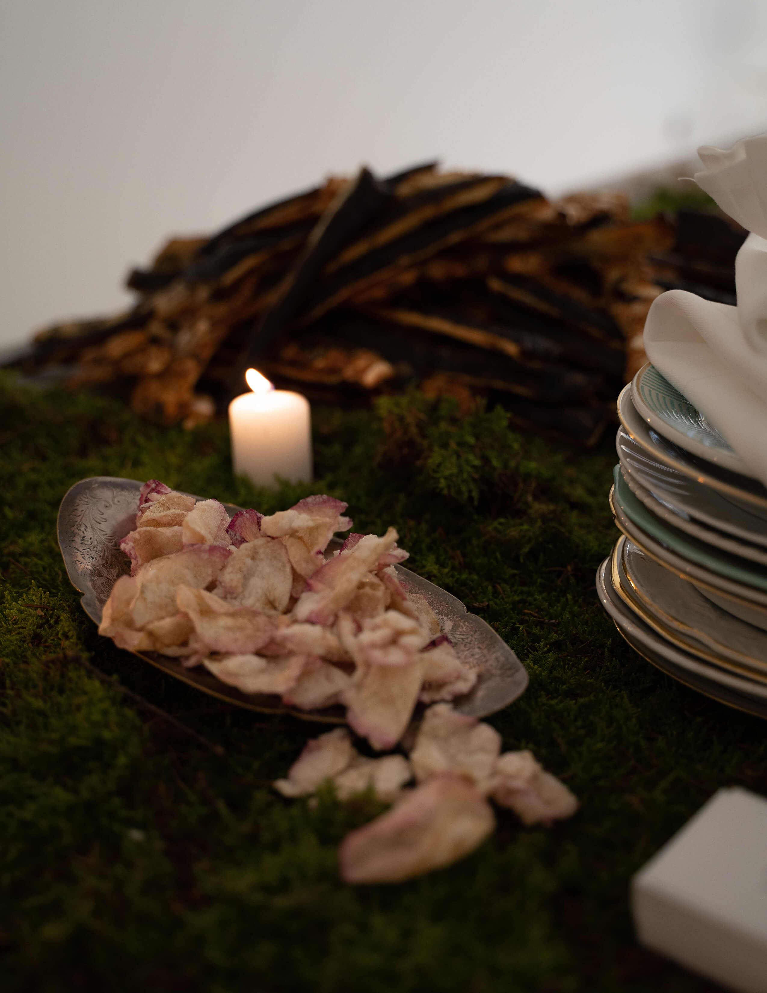 A plate of something that looks like crispy pedals next to a candle and a pile of ceramic plates on the moss's surface.