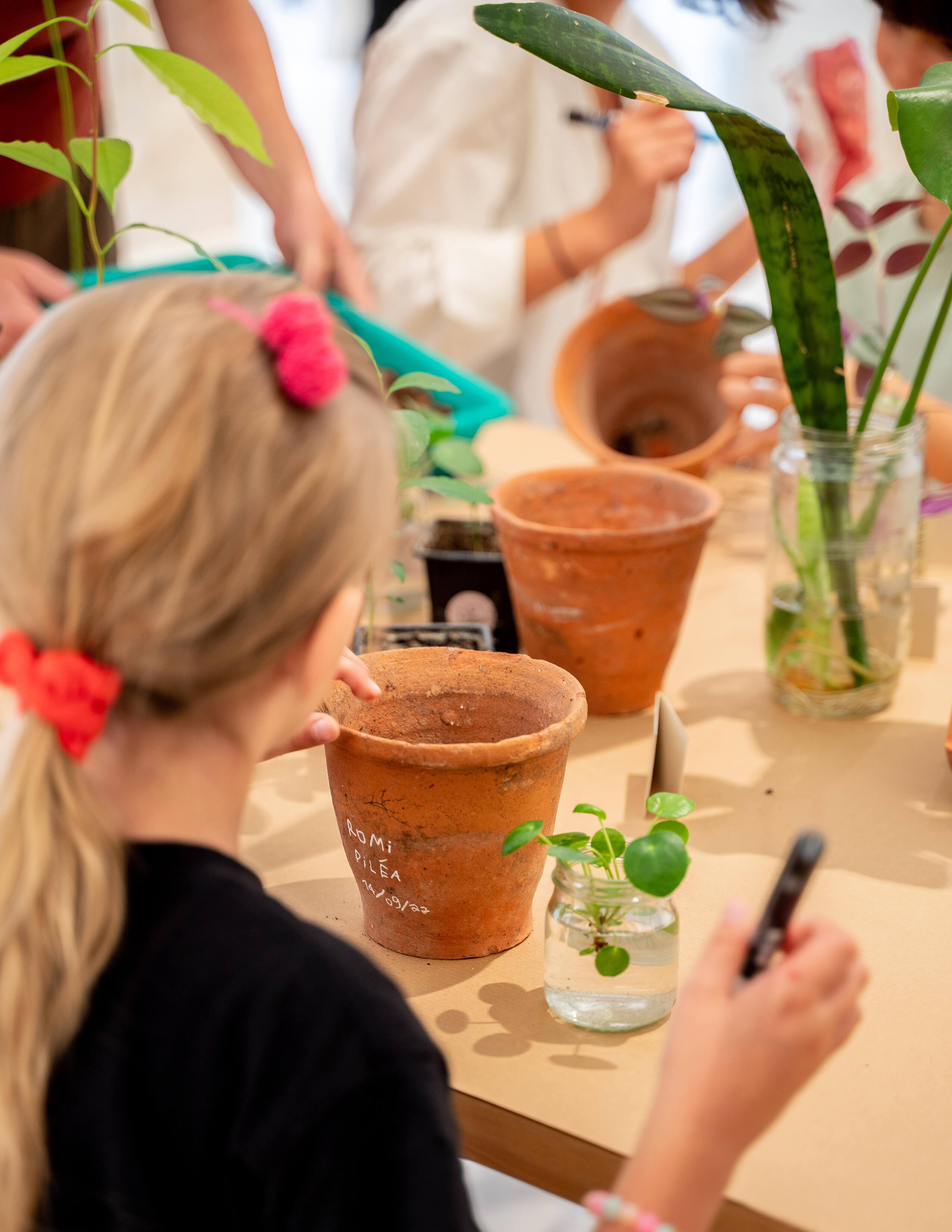 A kid with a red hair-tie holding a pen in her hand is white terracotta pots and plants are growing in glass bottles on the table.