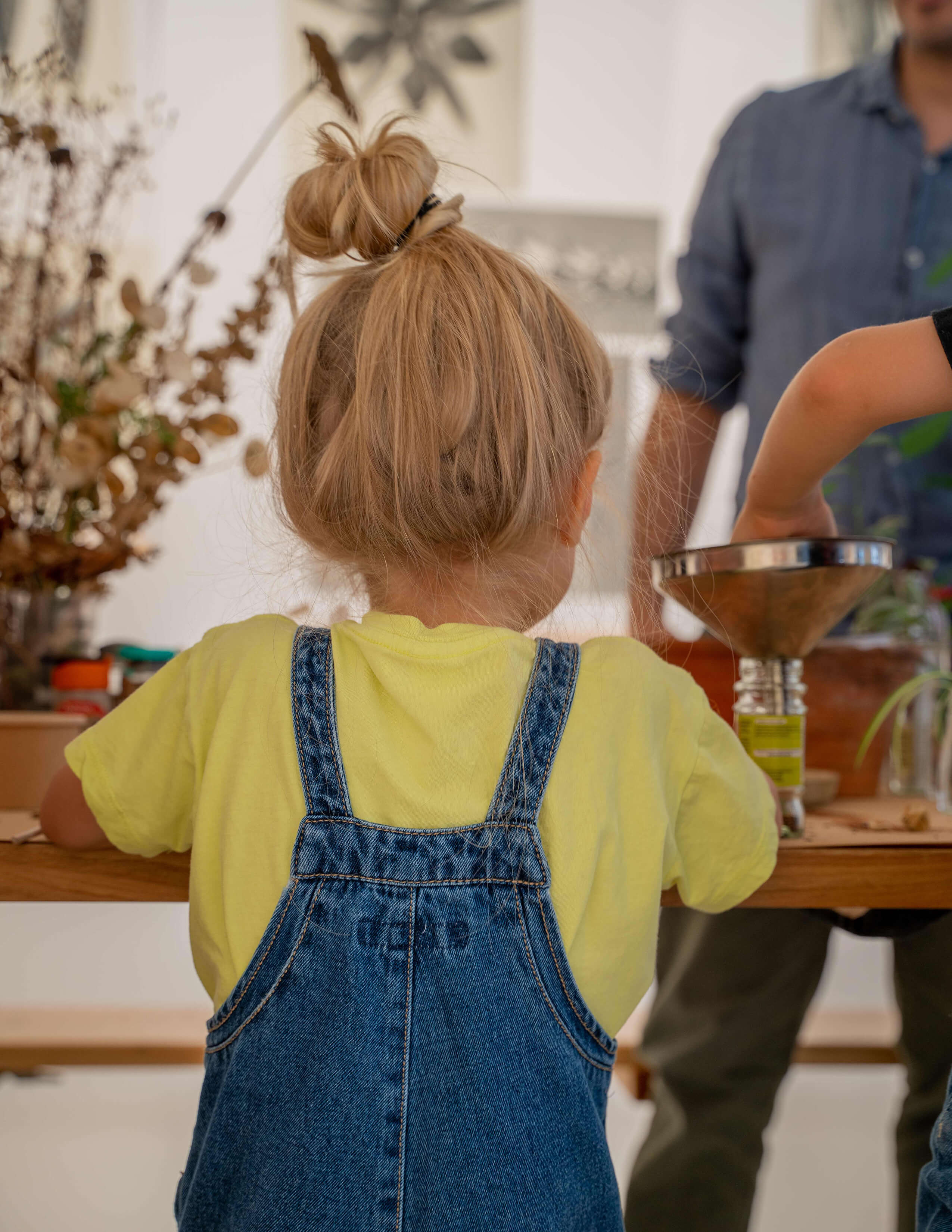 A kid with blonde tied-up bun hair wore a yellow jumper underneath the denim dungarees looking at people working on the table.