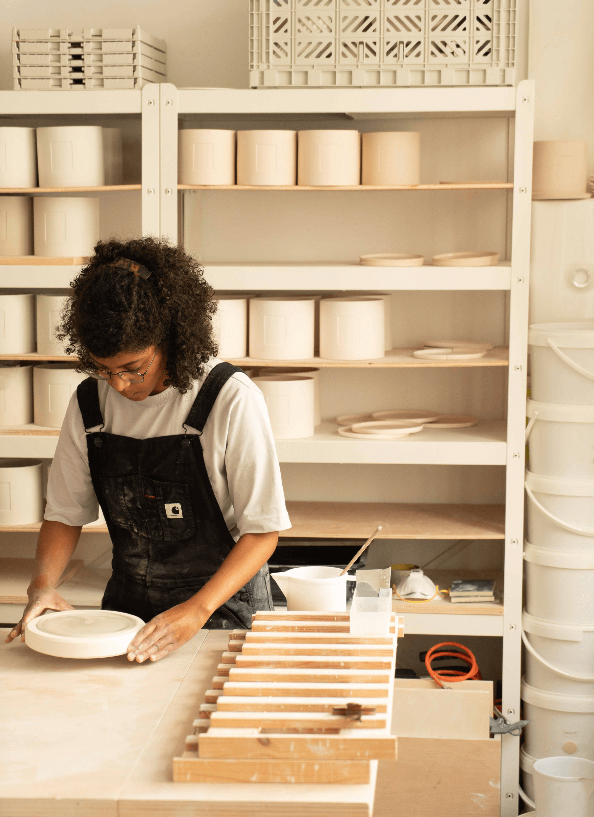 Salima adjusting the project that she is making on the table and she is wearing a cream t-shirt with a black dungarees. There are two wooden crafts on the table that are used to separate undried ceramic projects. Behind Salima, there is a shelf of ceramic pots stocked on the shelf with some ceramic saucers as well.