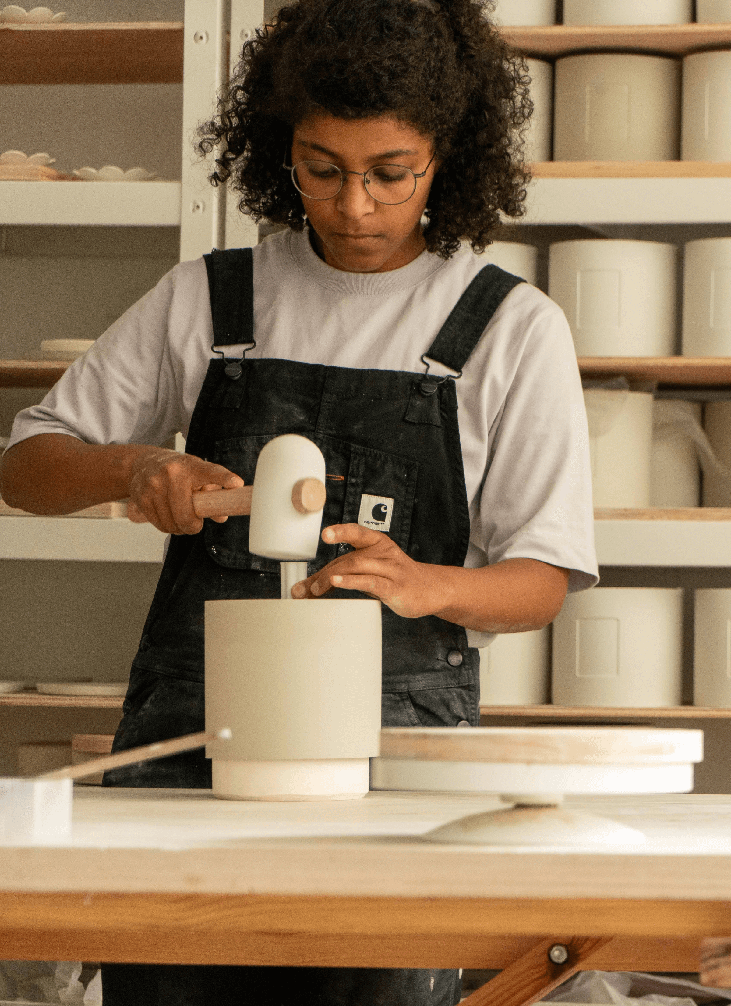 Salima used a hammer to label a symbol at the bottom of the ceramic pot that she's making on the table. She's wearing a cream t-shirt with a black dungarees and in the background, there are a full shelf of ceramic pots and flower shape of germination discs.