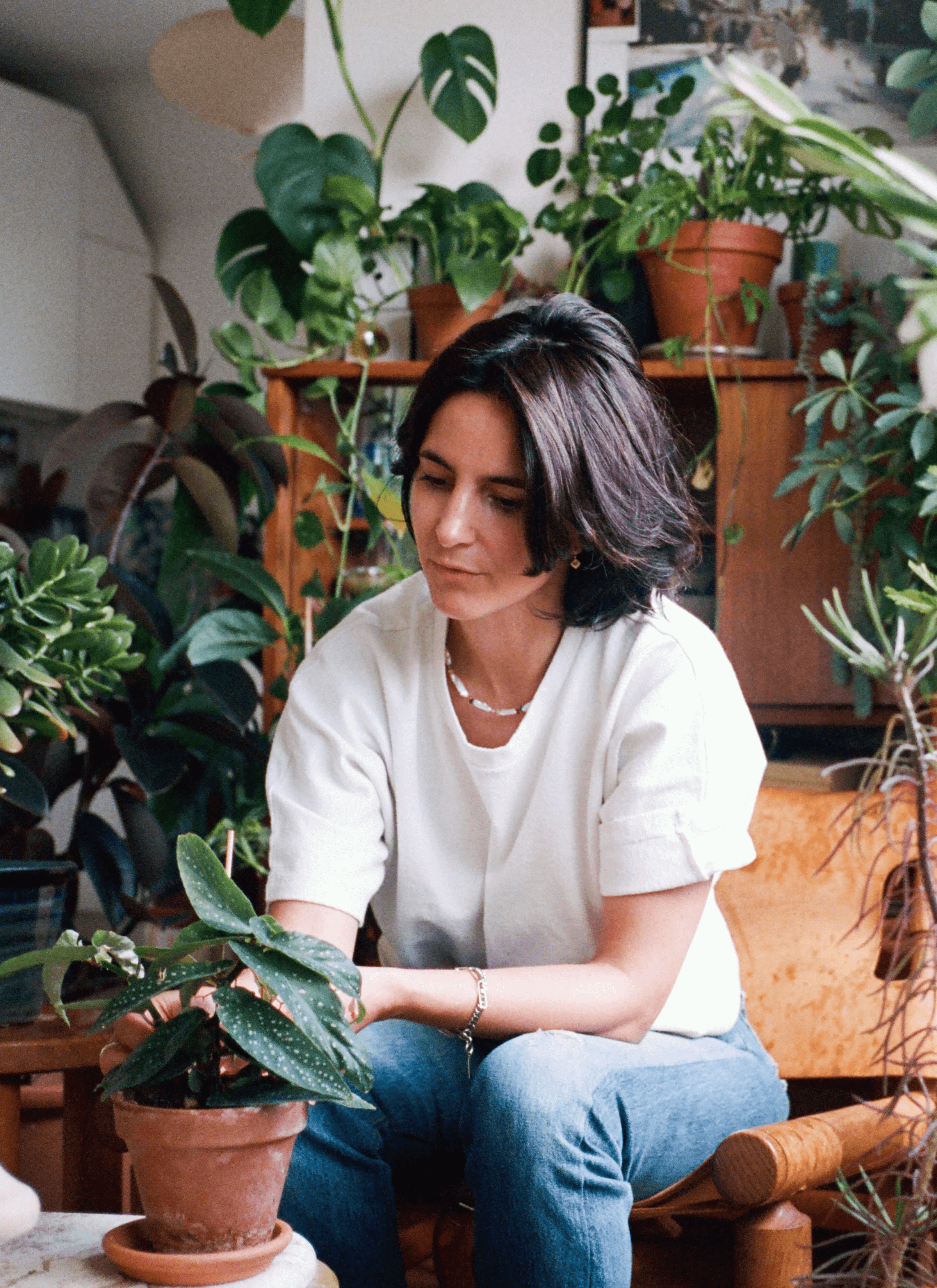 Leslie David is sitting on the chair taking care of the Angel wing begonia plant on a white marble table in her studio, surrounded by a collection of house plants.