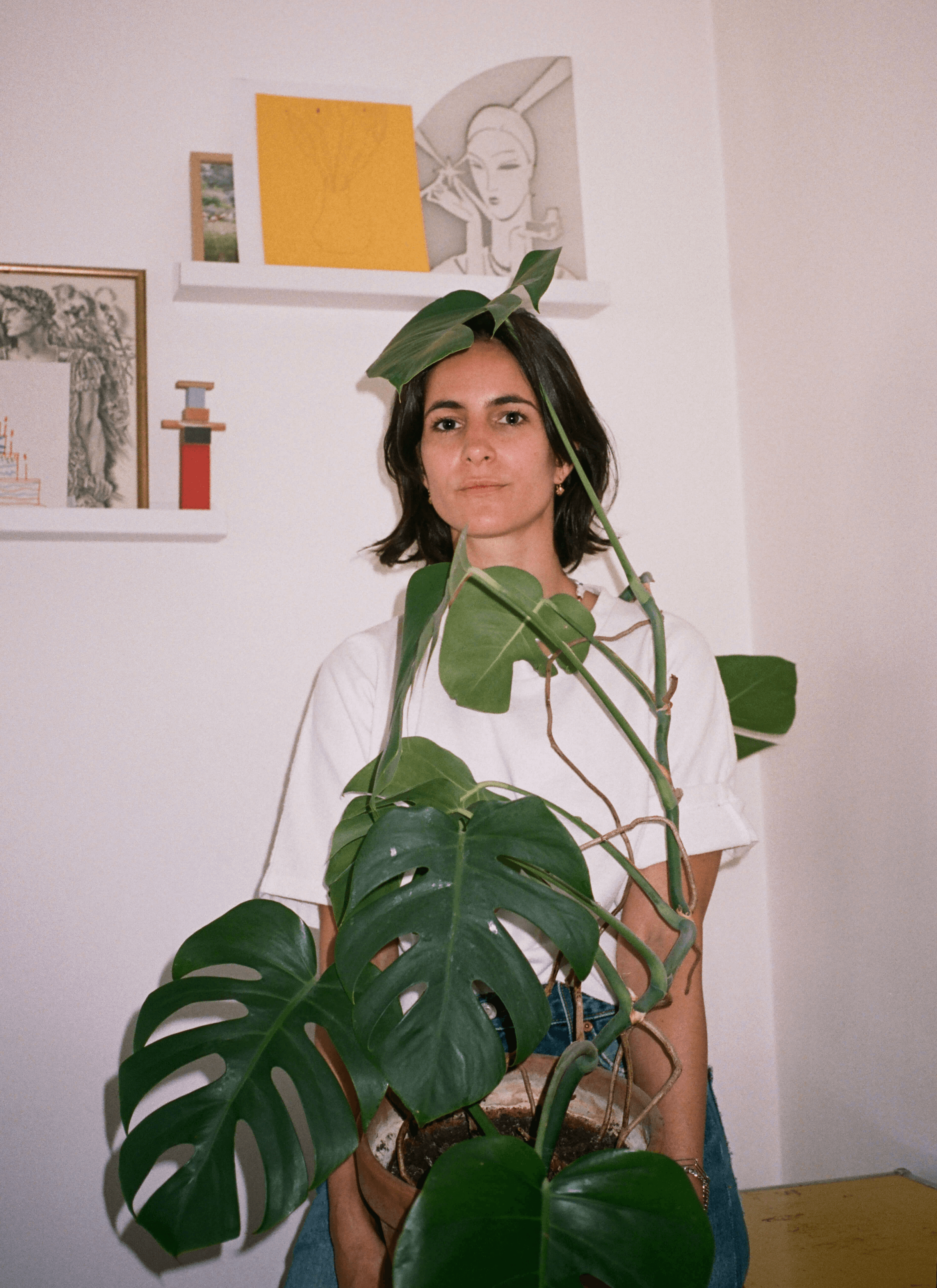 Leslie David is holding a pot of Monstera in front of her and there are some paintings, and art on the white wall shelf.